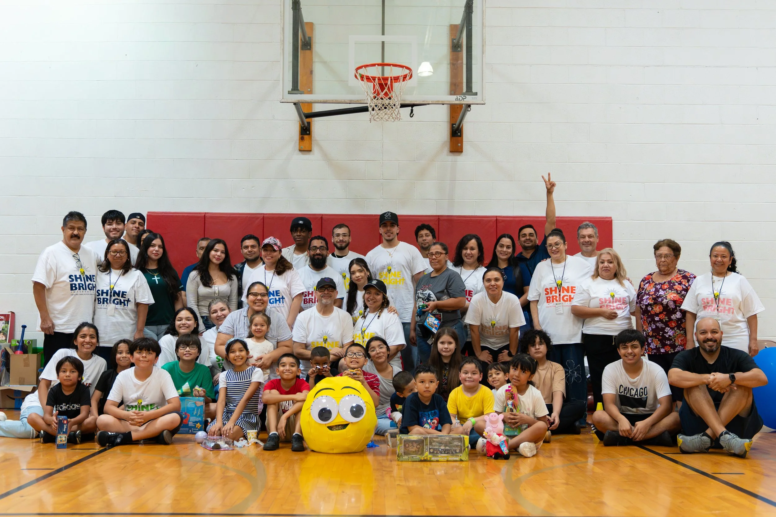 A large group of diverse people of all ages gathered inside a gym, posing for a group photo in front of a basketball hoop. The group includes children, adults, and seniors, some wearing shirts with the slogan 'Shine Bright.' There are balloons and toys on the floor, and the background features a plain white wall.