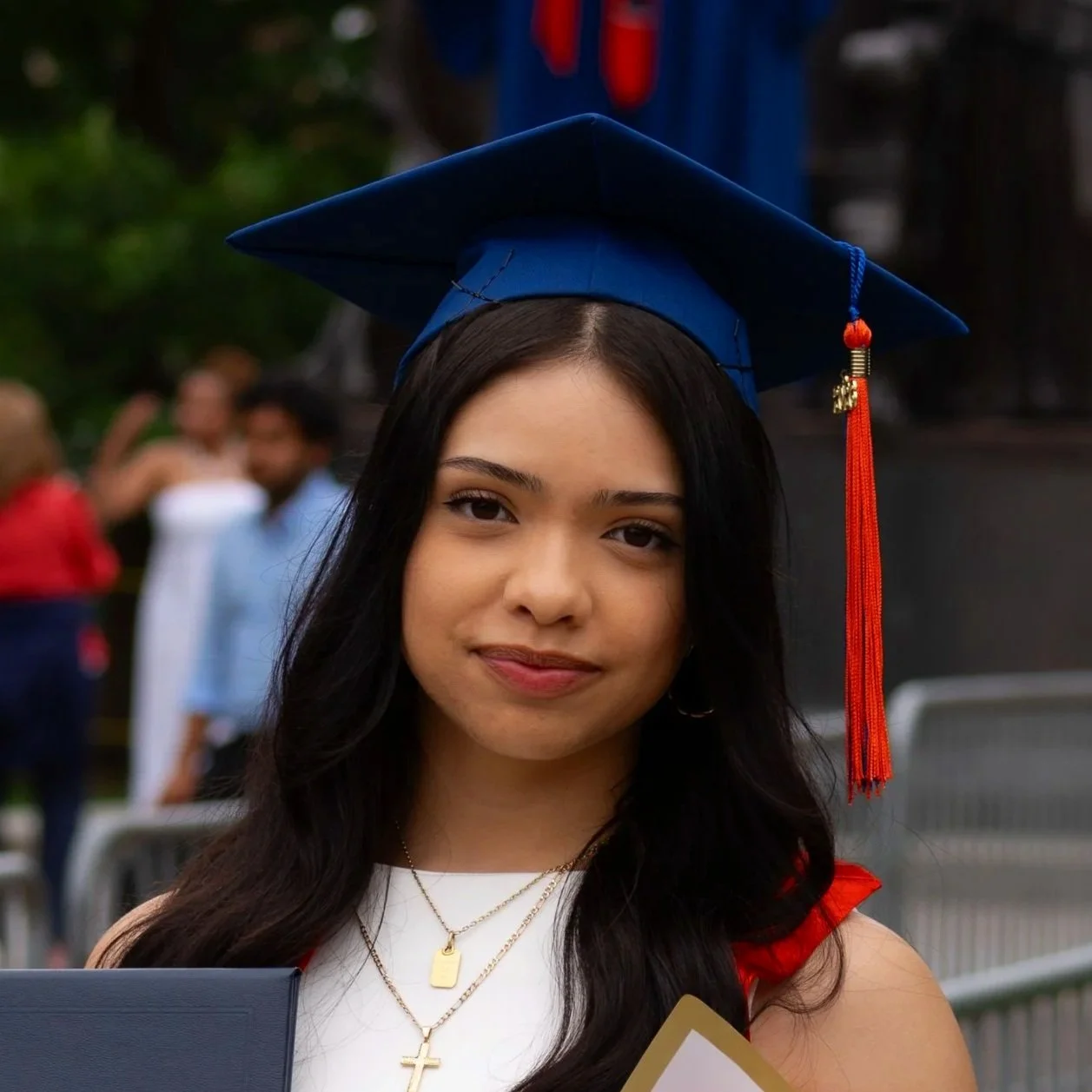 A young woman wearing a blue graduation cap with a red tassel, standing outdoors, with other people blurred in the background.