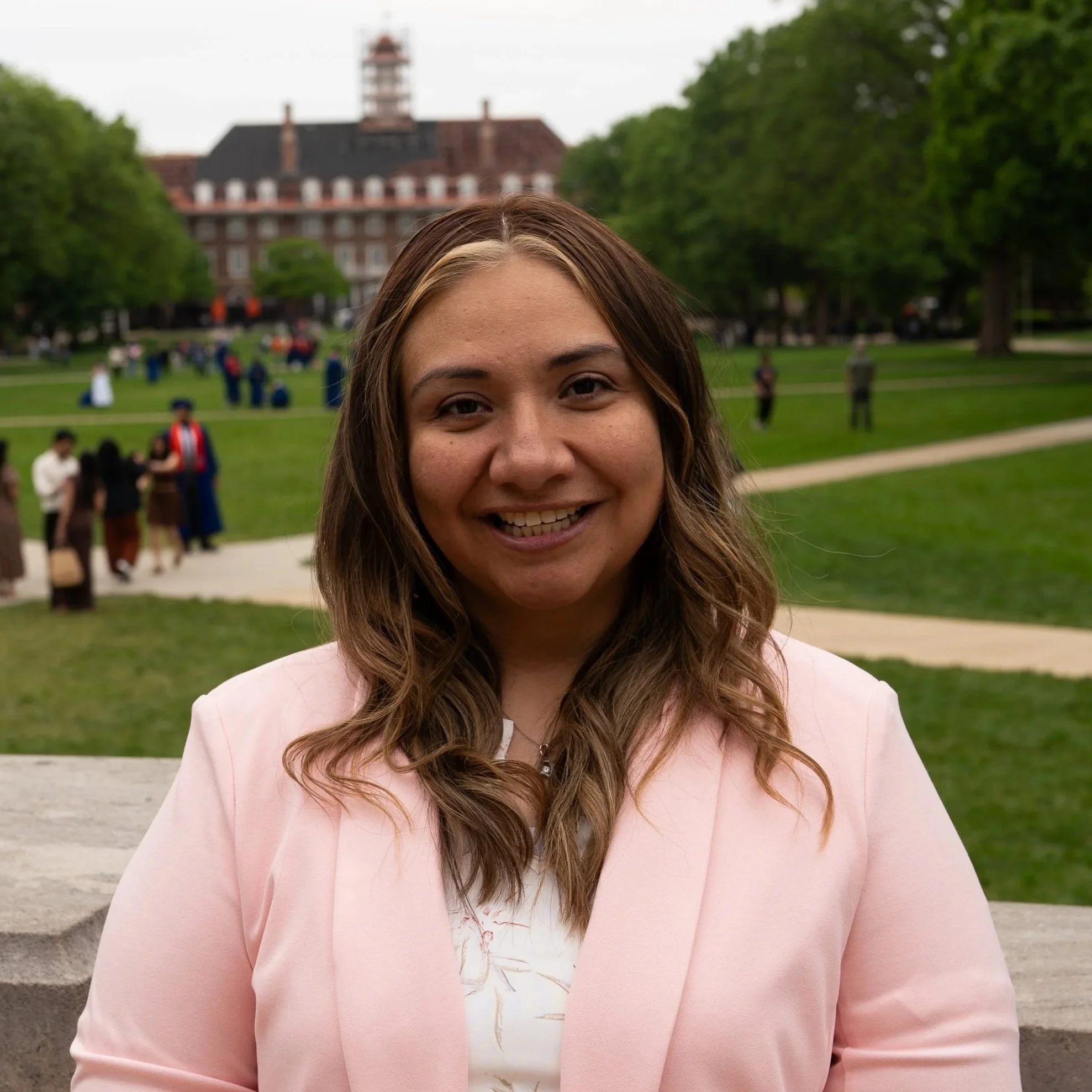 A woman with long, wavy brown hair smiling outdoors in front of a historic brick building with a clock tower, green trees, and a park with people in the background.