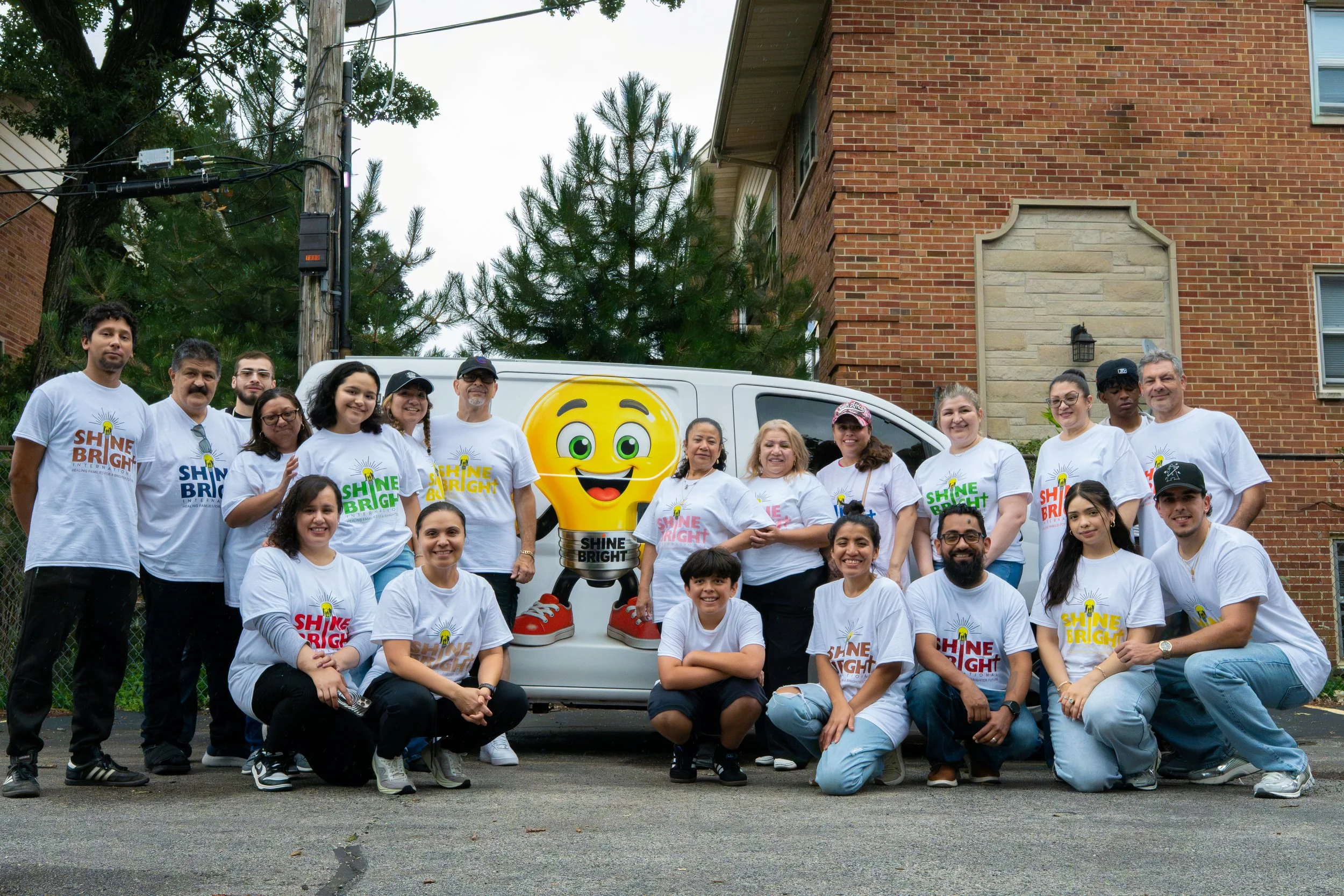 Group of diverse people gathered outdoors in front of a mural featuring a large smiling lightbulb mascot with red sneakers. The group is wearing white t-shirts with the phrase 'Shine Bright' and a lightbulb logo, smiling for the camera.
