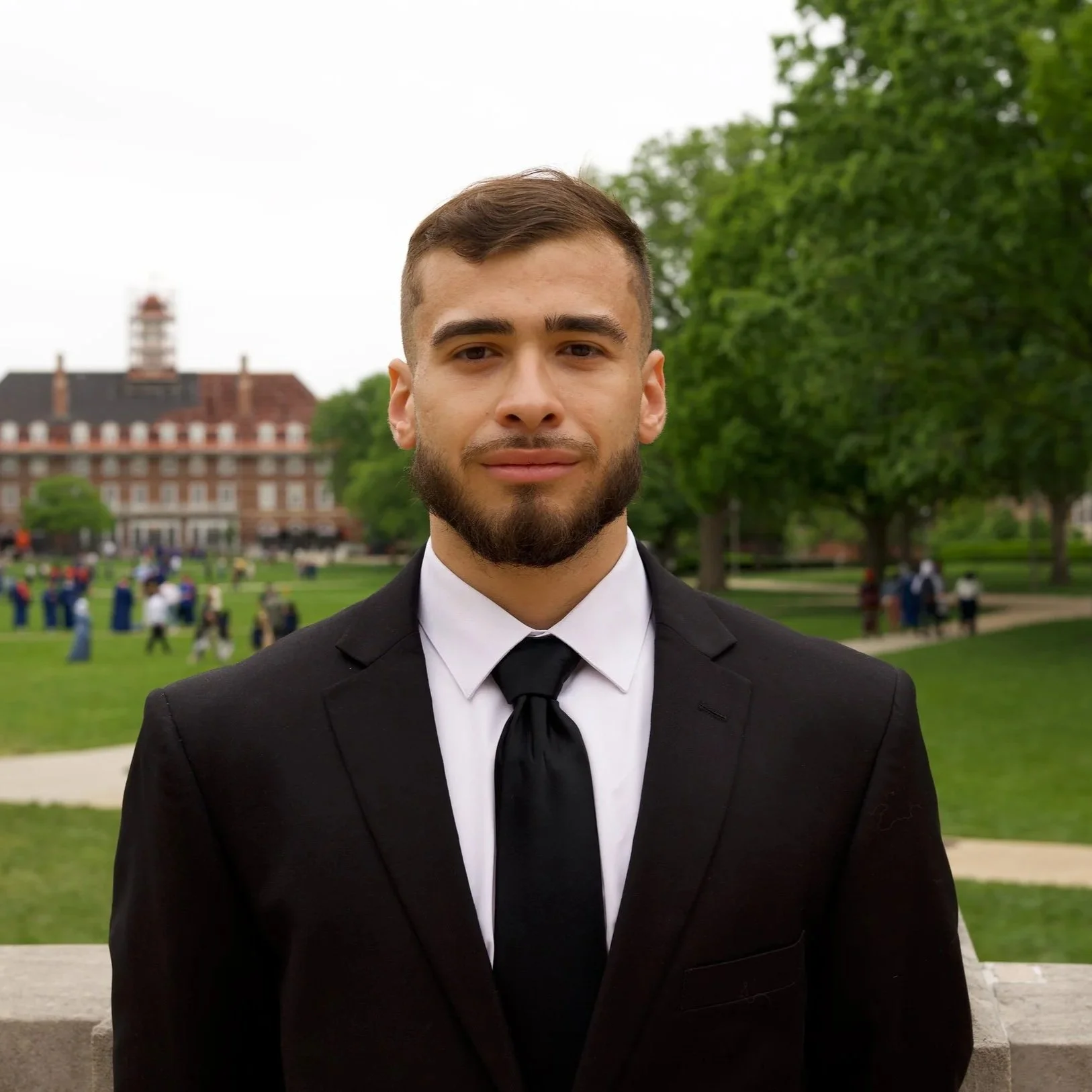 Young man in a black tuxedo and tie standing outdoors in front of green trees with a large building in the background.