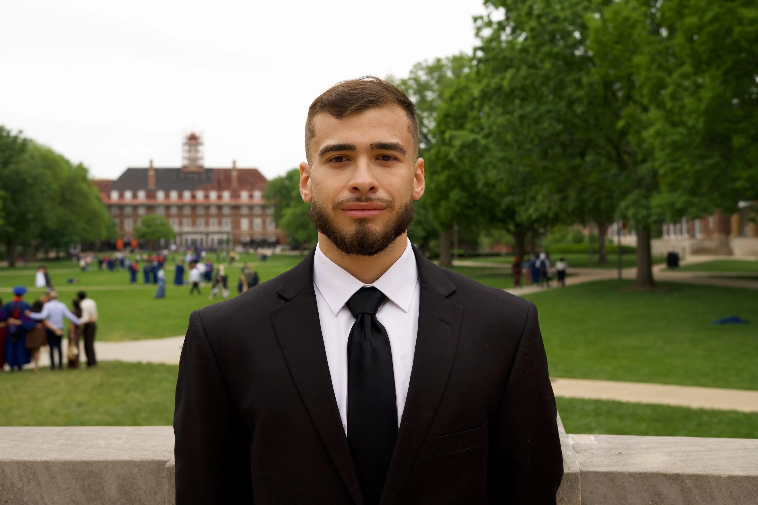 A young man with brown hair and a beard wearing a black suit, white shirt, and black tie, standing outdoors on a park bench with green trees and a brick building in the background.