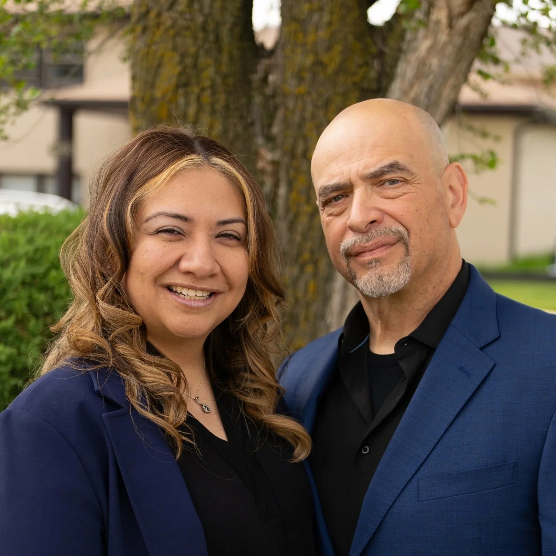 Close-up of a smiling woman with long, wavy light brown hair and a man with a bald head, goatee, and mustache, both wearing blue suits, standing outdoors in front of a large tree with a brown bark trunk and green foliage.