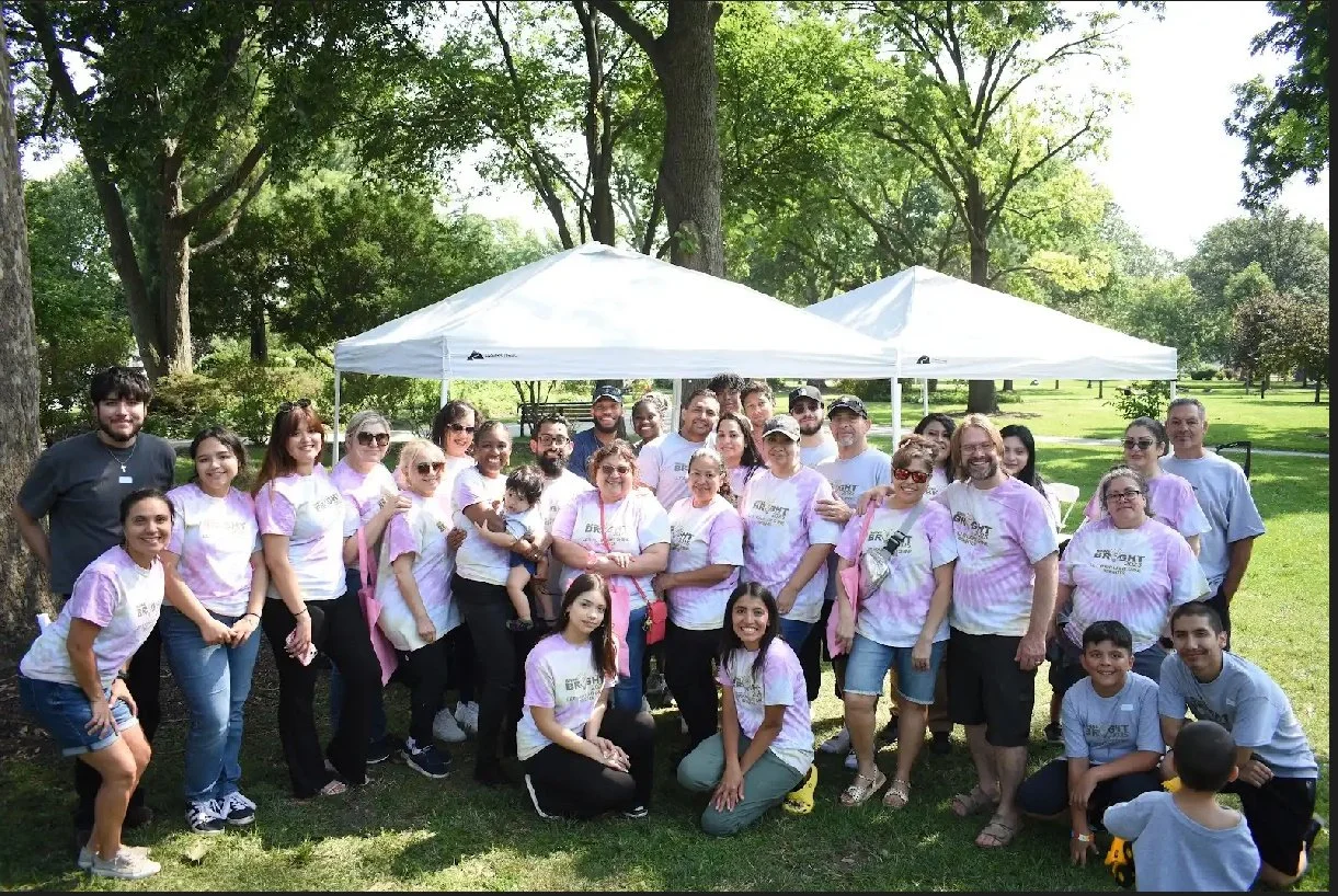 Group of people gathered outdoors in a park with trees, standing under two white canopies, smiling for a photo.