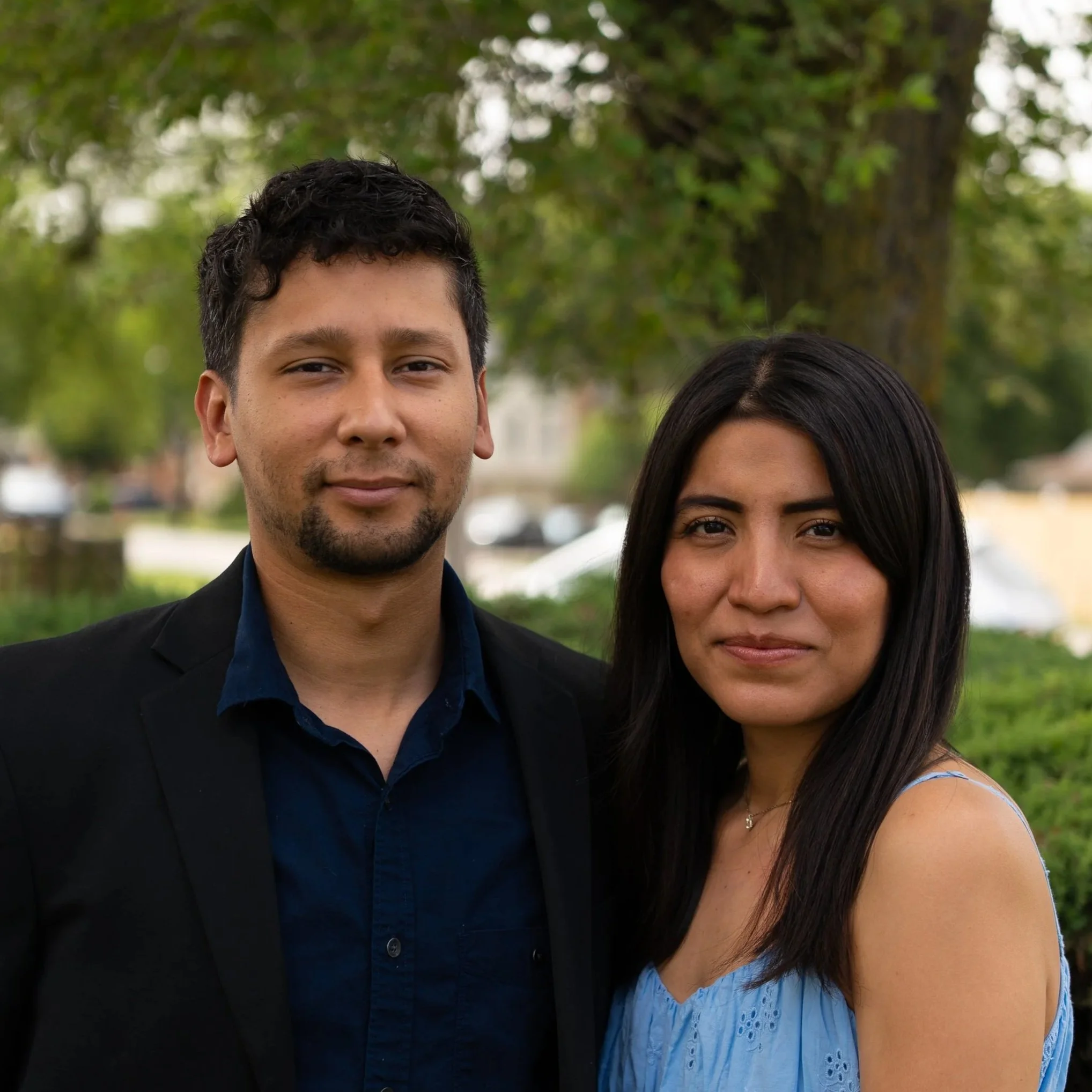 Close-up of a man and woman standing outdoors with trees in the background.