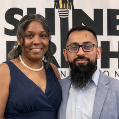 A woman and a man posing together at an event, standing in front of a backdrop with partial text and logo.