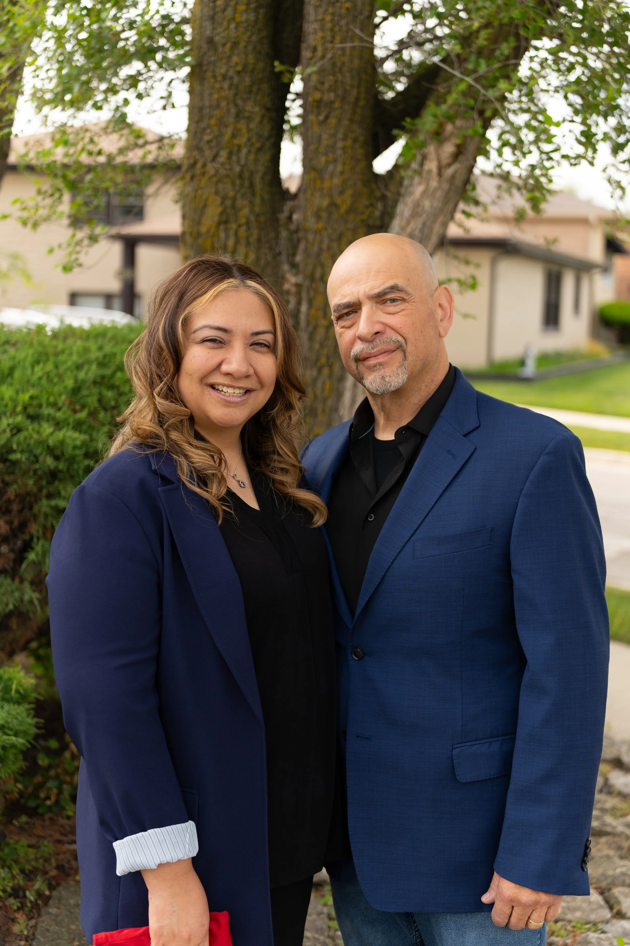 A smiling woman and an older man standing outdoors in front of a large tree, house, and greenery on a partly cloudy day.