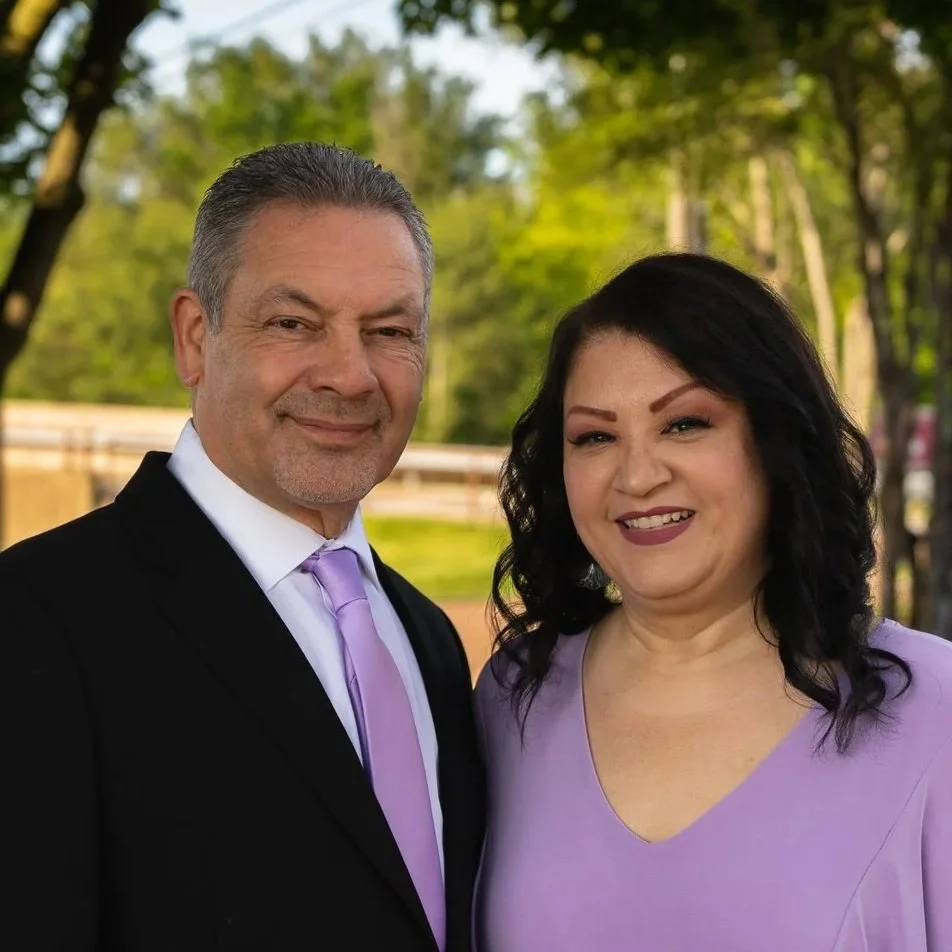 A man and woman standing outdoors, smiling, with trees and a fence in the background. The man is wearing a black suit with a white shirt and a lavender tie, while the woman is dressed in a lavender top.