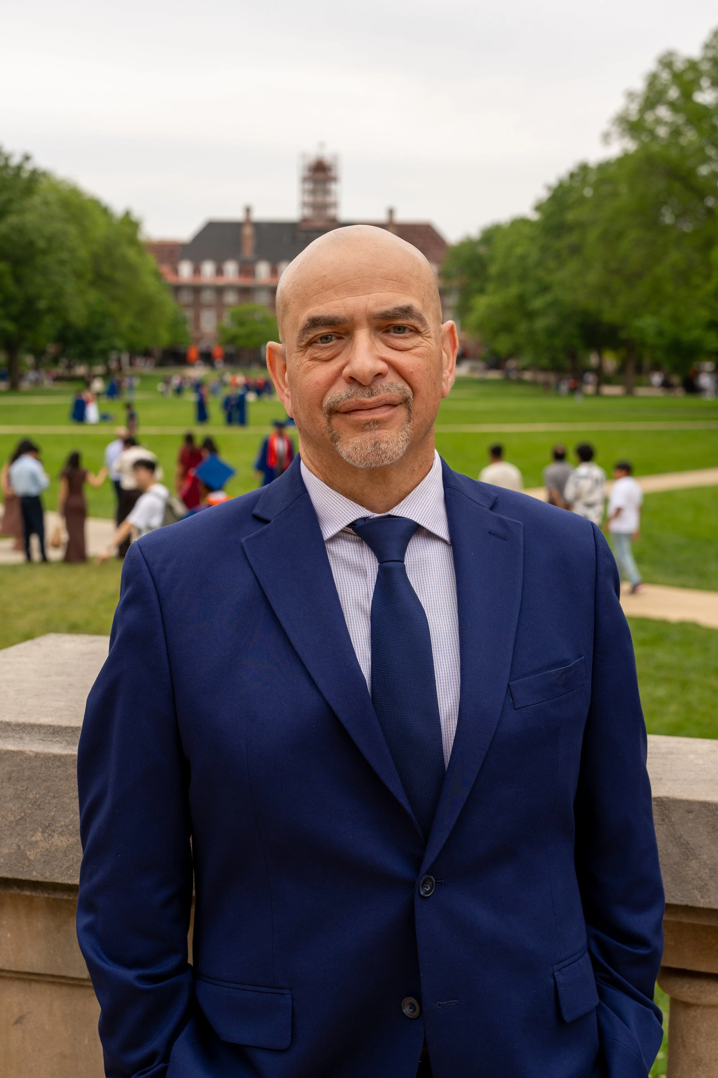 A man in a navy blue suit and tie posing outdoors in front of a park with green trees and a historic building with a clock tower in the background.