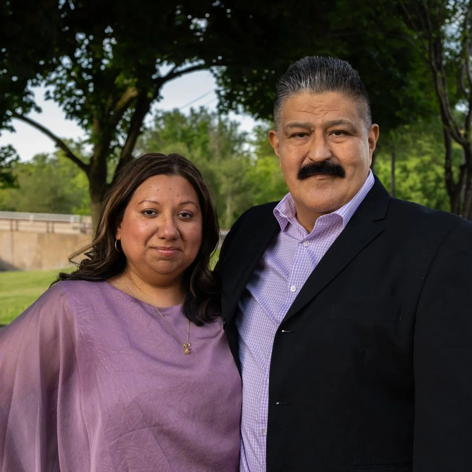 A man and woman standing outdoors, close together, smiling at the camera. The woman is wearing a purple dress and a gold necklace, and the man is wearing a black suit jacket with a purple checkered shirt. They are under a tree in a park or garden, with green leaves and a stone wall in the background.