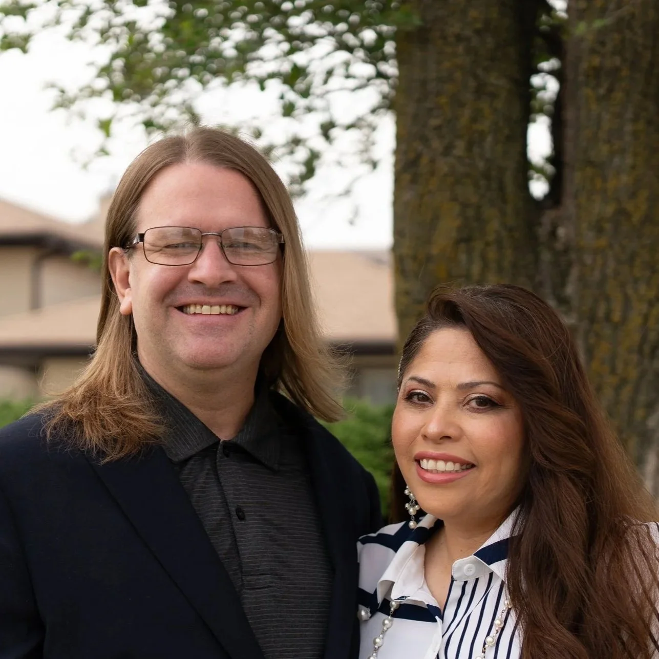 A man with long blond hair, glasses, and a black suit, standing next to a woman with long dark hair, wearing a striped shirt, both smiling outdoors near a large tree.