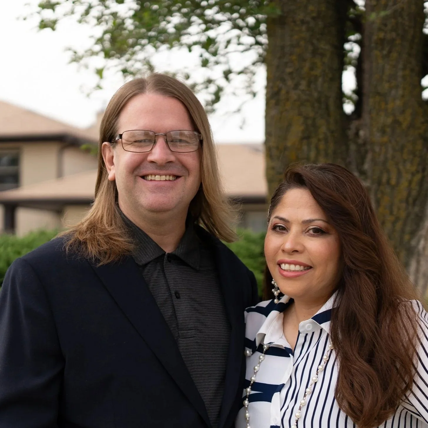 A smiling man with long blonde hair, glasses, wearing a black jacket, standing next to a smiling woman with long brown hair, wearing a white and navy striped blouse, outdoors near a tree with buildings in the background.