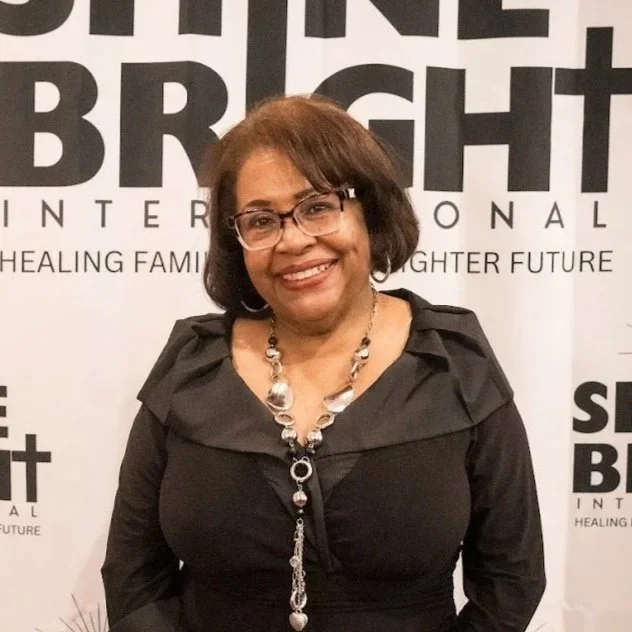 Smiling woman with glasses and short hair, wearing a black jacket and necklace, standing in front of a banner that says "Bright International"