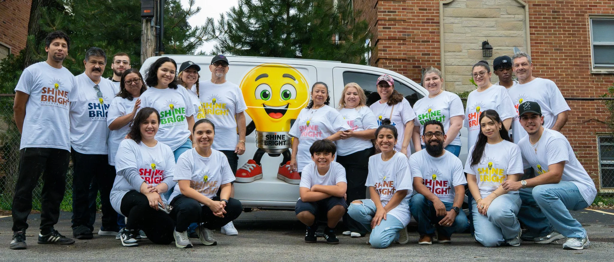 A diverse group of people, including children and adults, standing outdoors in front of a white van with a cartoon light bulb mascot. They are smiling and wearing matching white T-shirts with the words 'Shine Bright' in colorful letters, participating in a community event.