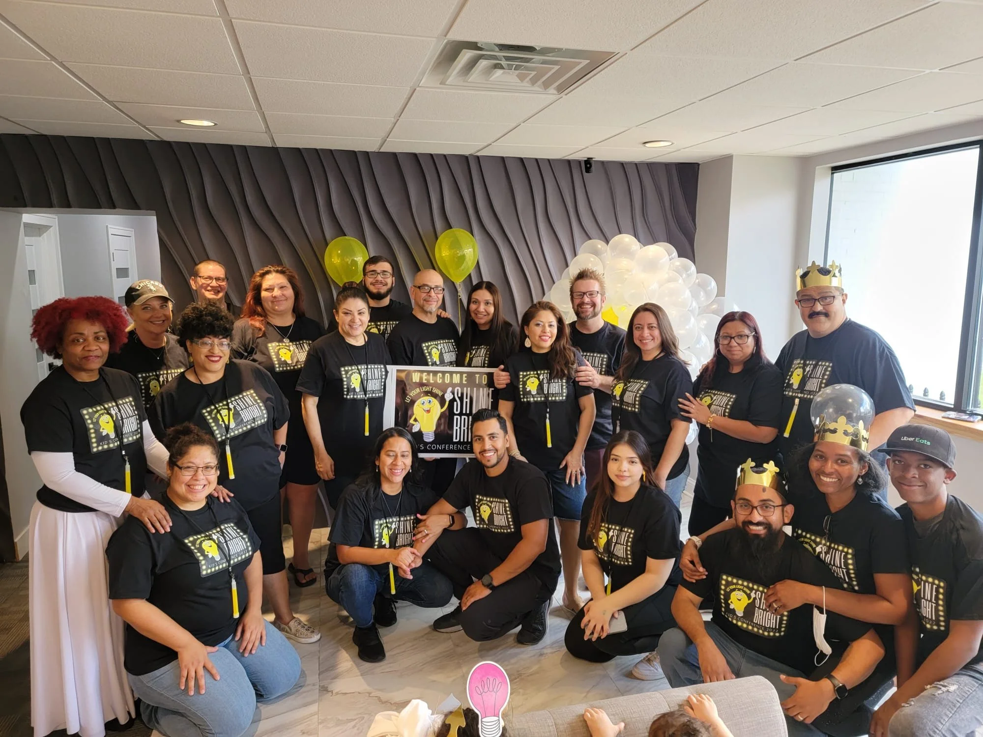A group of people gathered indoors for a celebration, wearing matching black t-shirts with a yellow lightbulb graphic and text, with balloons and signs in the background.