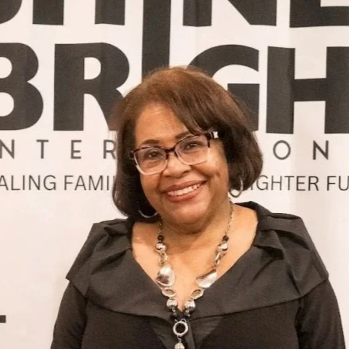 An older African American woman with shoulder-length hair and glasses, smiling, wearing a black dress and a statement necklace, standing in front of a white wall with black text.