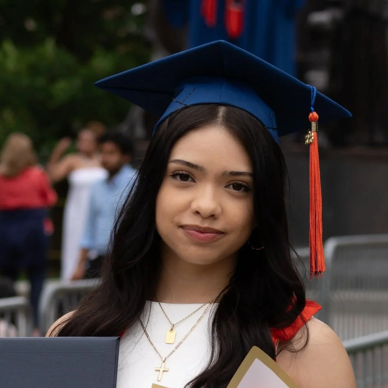 Young woman with long dark hair wearing a blue graduation cap with red tassel, wearing gold necklaces including a cross, smiling at graduation ceremony.