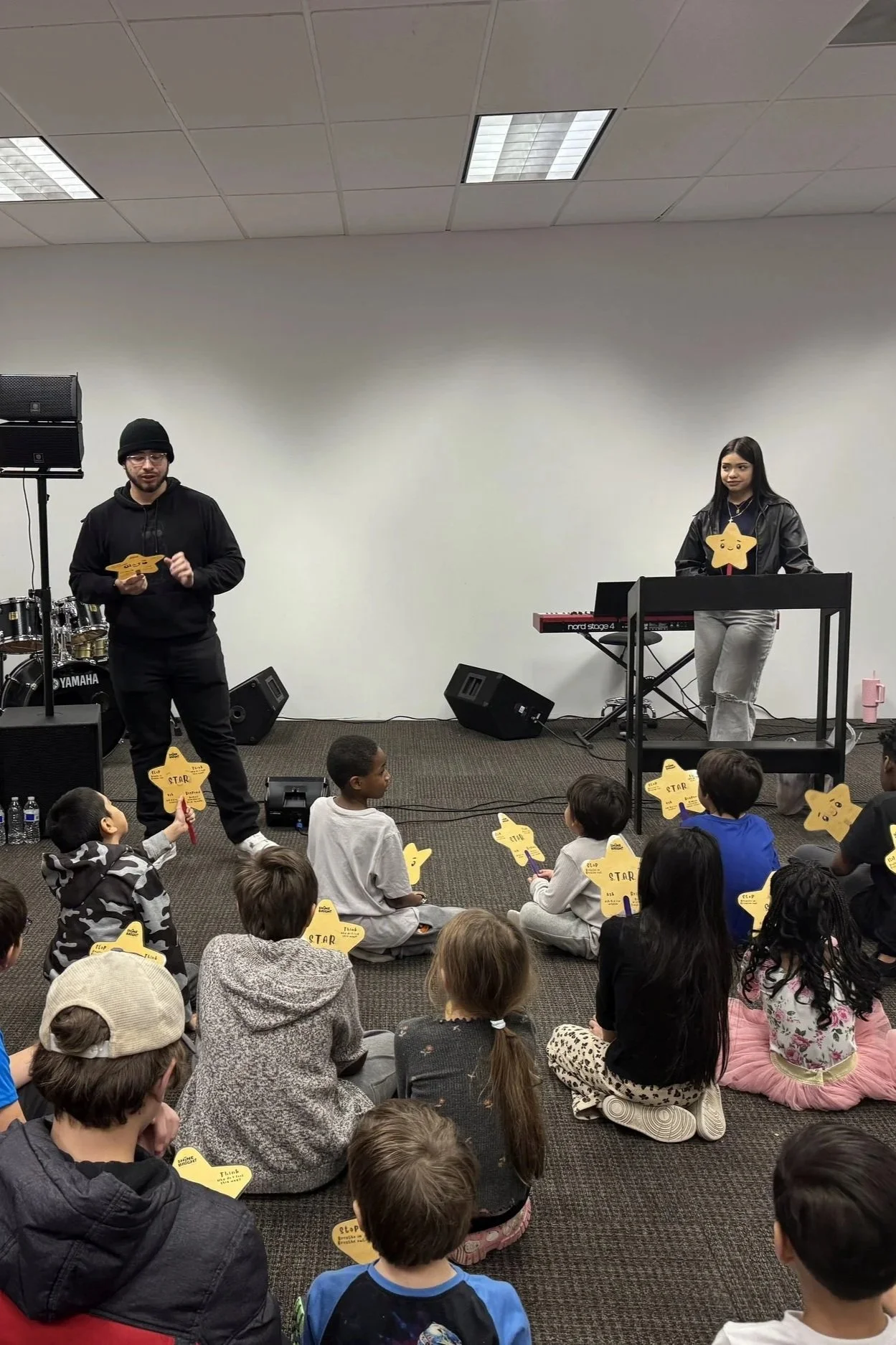 Children attending a performance or presentation in a room with adults, with some holding yellow star-shaped cards that have writing on them, while a man and woman stand at the front, the woman behind a keyboard and the man holding a paper, in a room with white walls and ceiling lights.