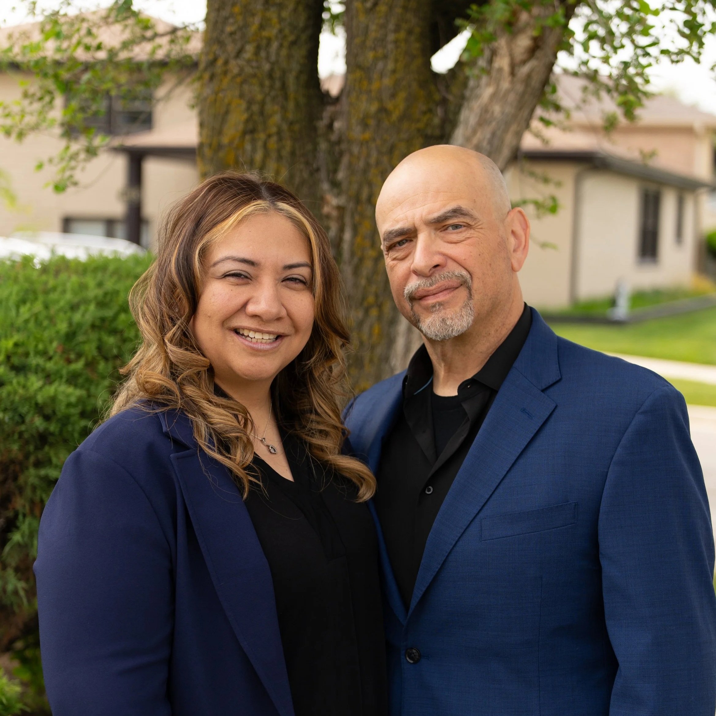 Close-up of a smiling young woman and an older man standing outdoors near a tree, with blurred buildings and greenery in the background.