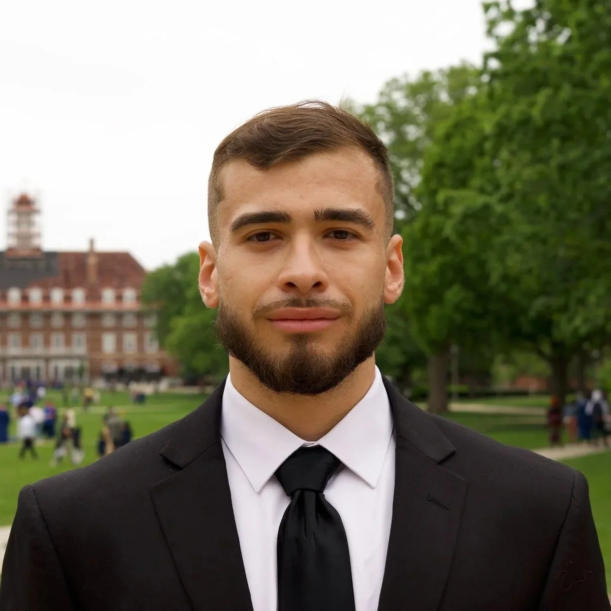 A young man in a black suit and tie standing outdoors on a cloudy day with green trees and a building in the background.