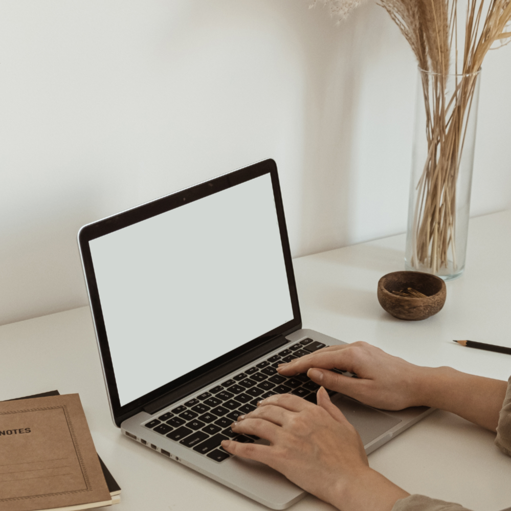 Person typing on a laptop at a white desk with a notebook, a black pen, and a glass vase with dried plants.