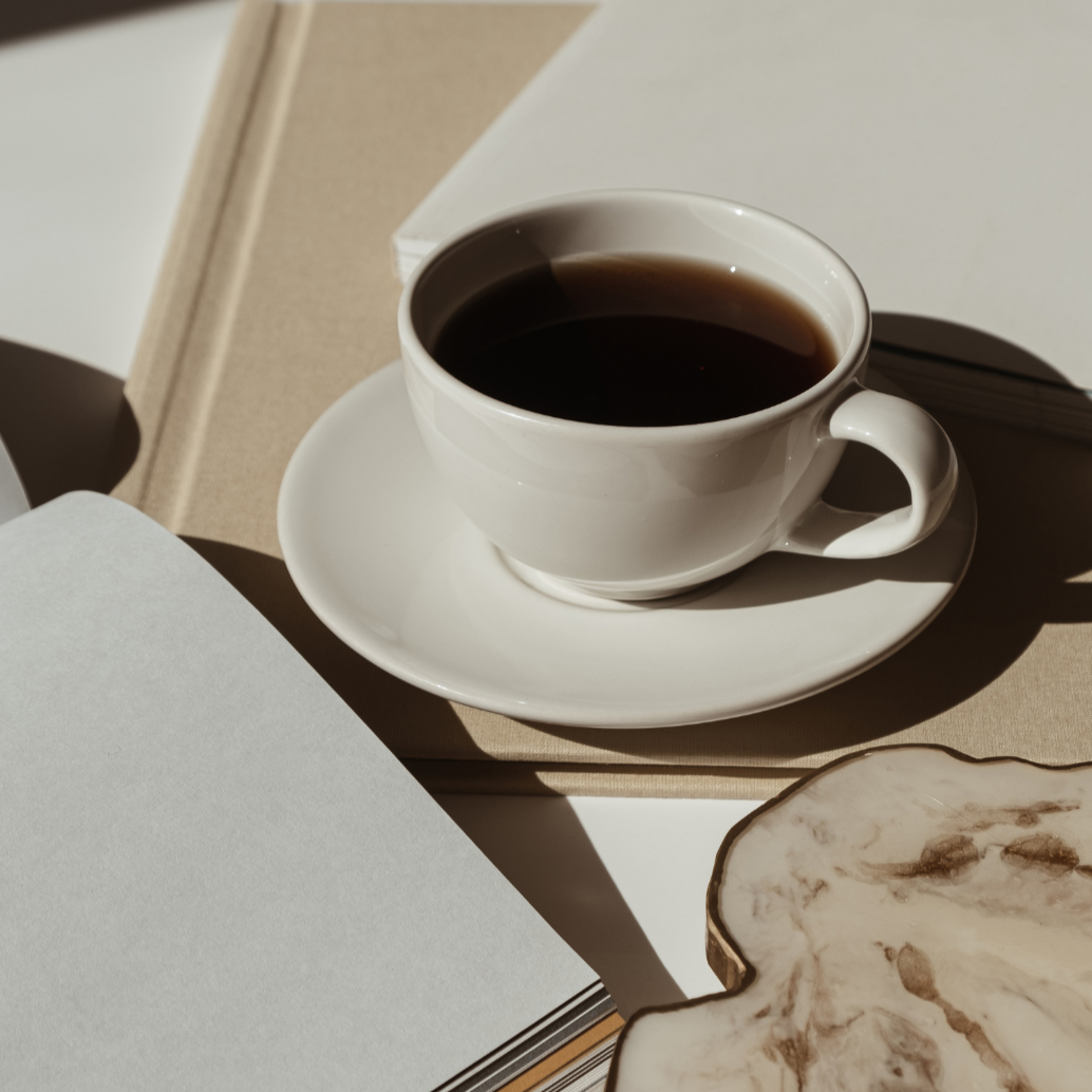 A white cup filled with black coffee on a saucer, placed on a table with open books and a marble-patterned object, with sunlight casting shadows.