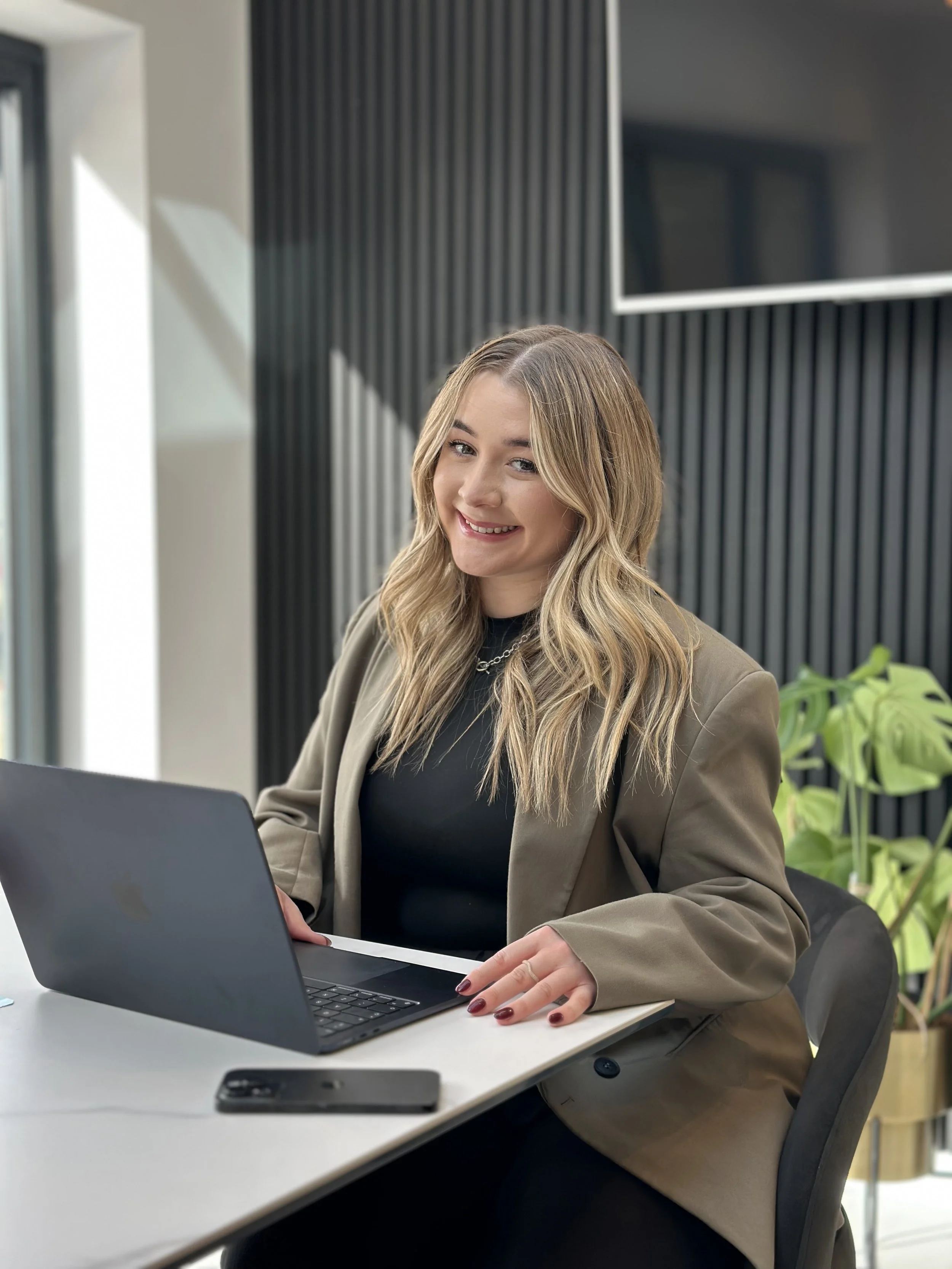 A young woman with long blonde hair sitting at a white desk working on a laptop, smiling, wearing a brown blazer and black top, in a modern office setting with a black textured wall and green plants in the background.