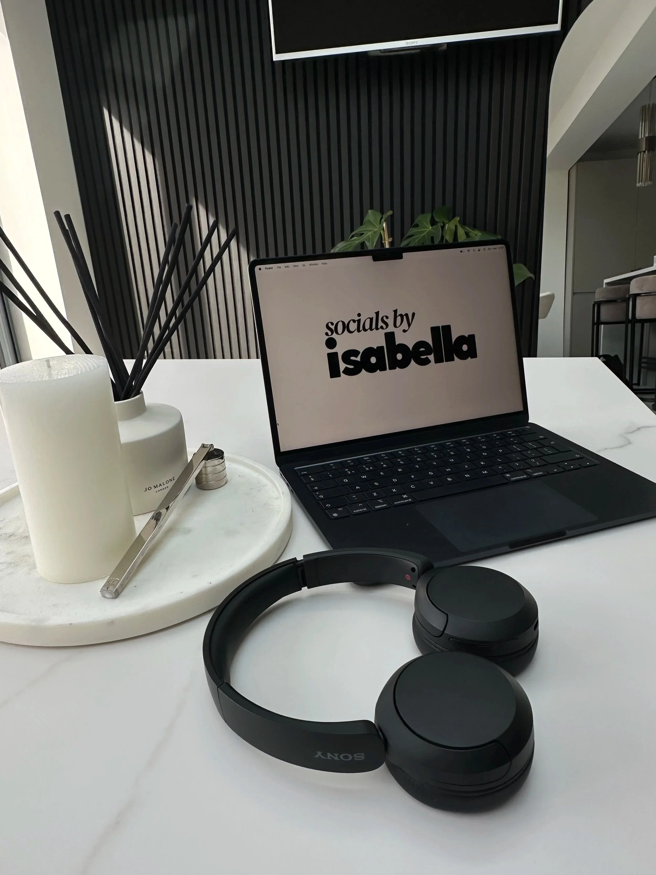Desk setup with Sony headphones, a laptop displaying 'socials by isabella,' a white tray with a candle, a pen, and a key, and a potted plant in the background.