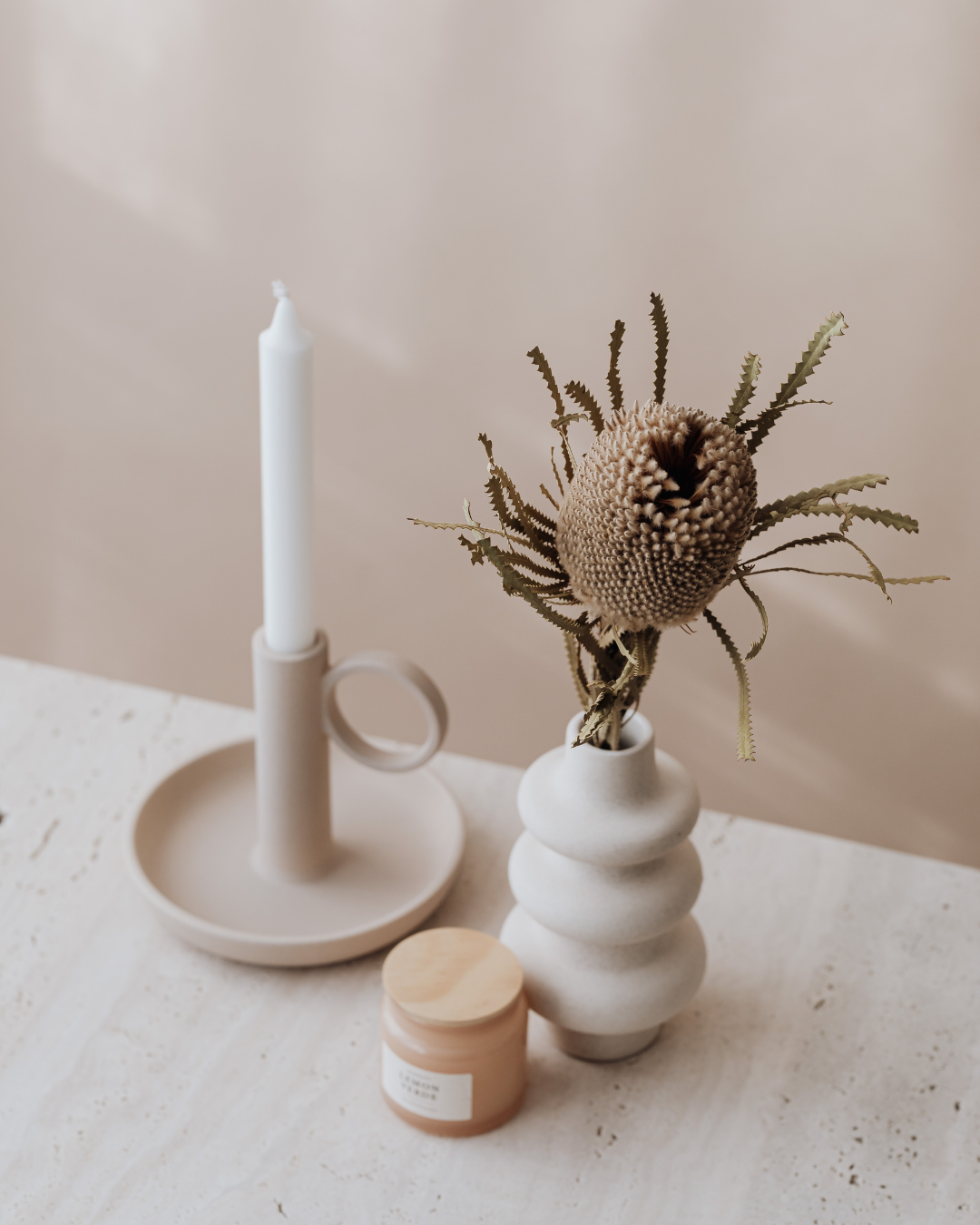 A white candlestick holder with a tall, unlit white candle, next to a beige vase holding dried protea flowers and fern leaves, and a small beige jar, all on a white textured surface against a plain light background.