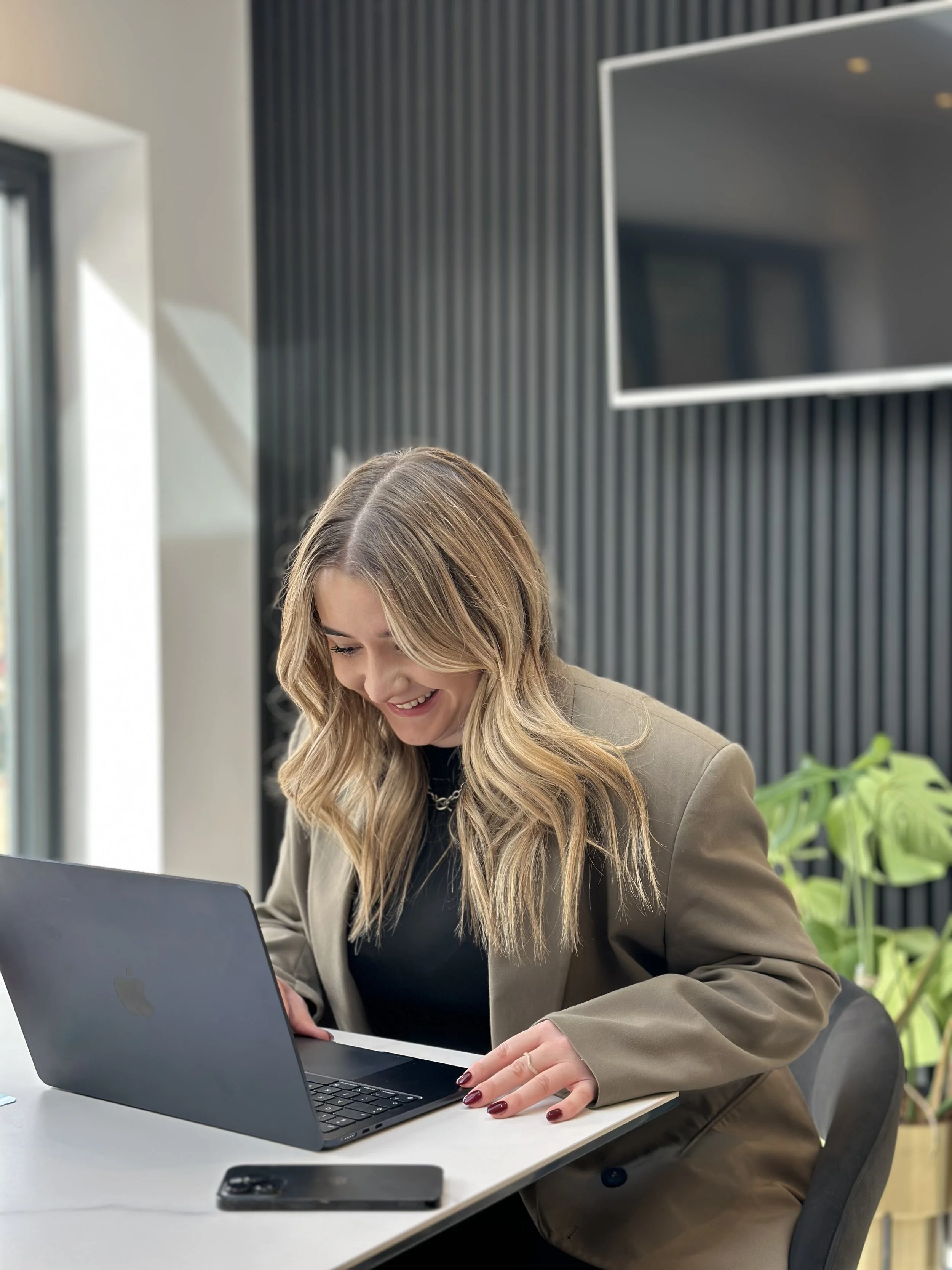 A woman with long blonde hair, smiling and working on a laptop at a white desk in a modern office with black walls, a large TV screen, and green plants.