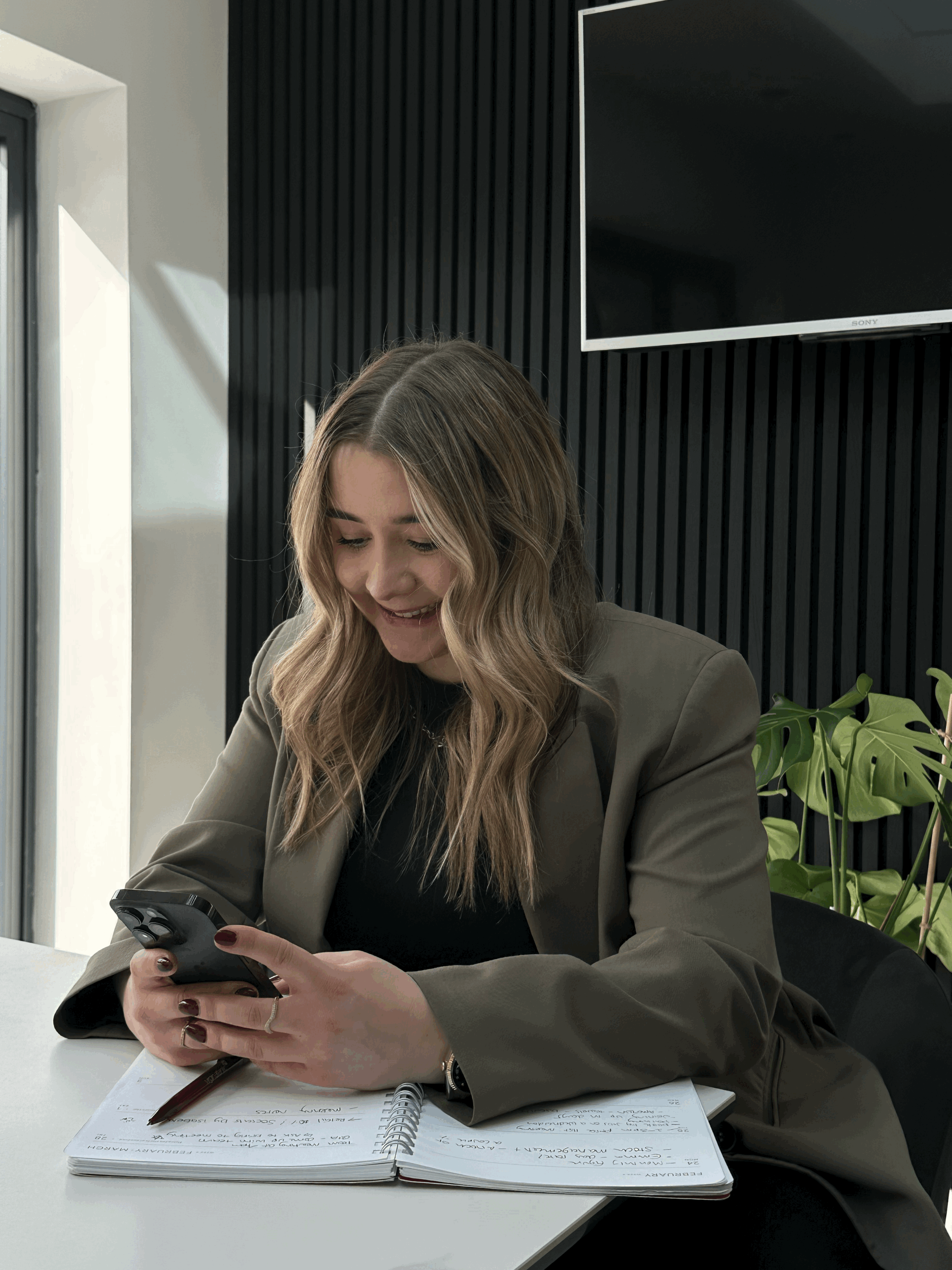 A woman with wavy blonde hair, wearing a brown blazer, is sitting at a white desk, smiling while looking at her phone. She has a notebook with handwritten notes and a pen in front of her. In the background, there is a black wall with vertical ridges, a large flat screen TV, and a green plant with large leaves.