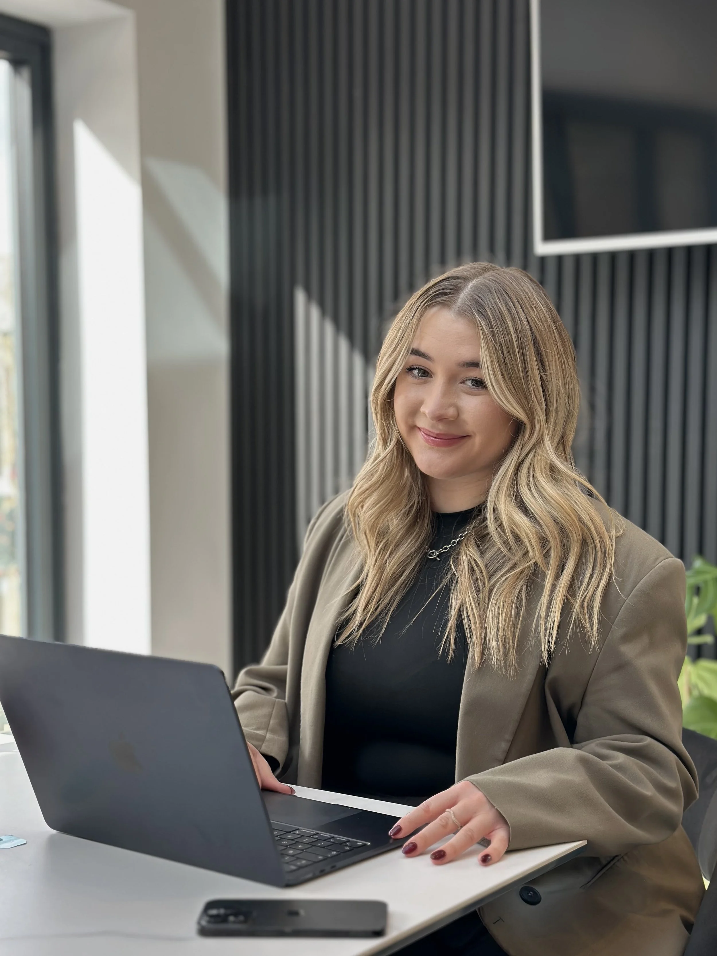 A woman with blonde wavy hair wearing a brown blazer and black top, working on a laptop at a desk in an office.