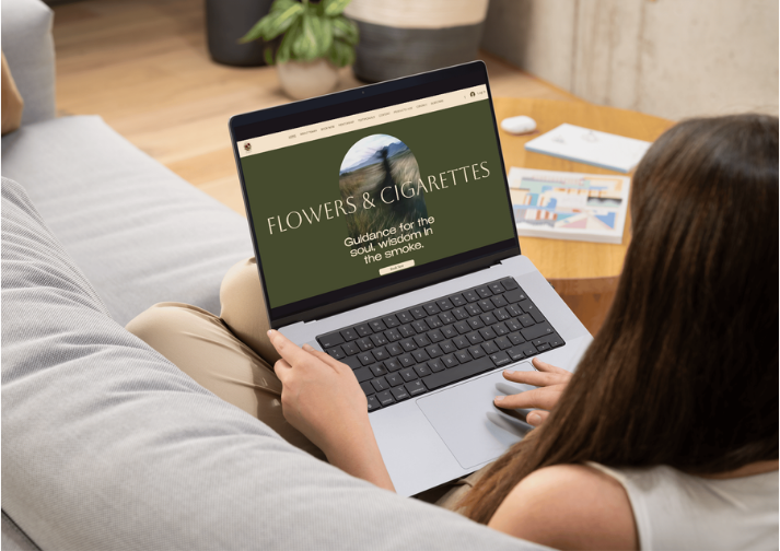 Person sitting on a gray sofa using a laptop with a website titled "Flowers & Cigarettes" on the screen, in a cozy room with a round wooden table and potted plants.