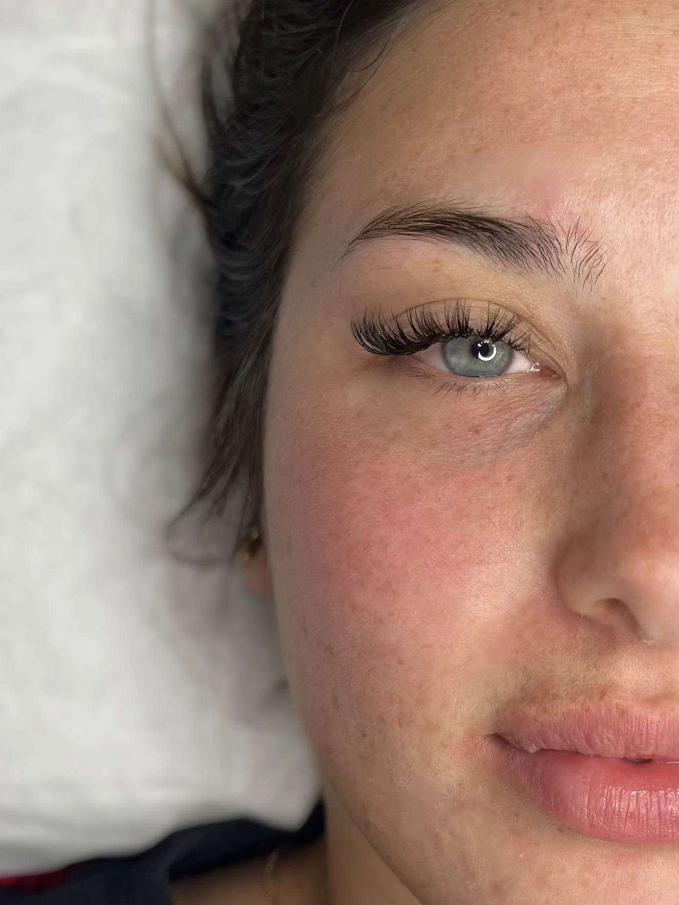 Close-up of a woman's face showing her eye with long dark eyelashes, arched eyebrow, and foundation-free skin with freckles and a slight blush on her cheek.