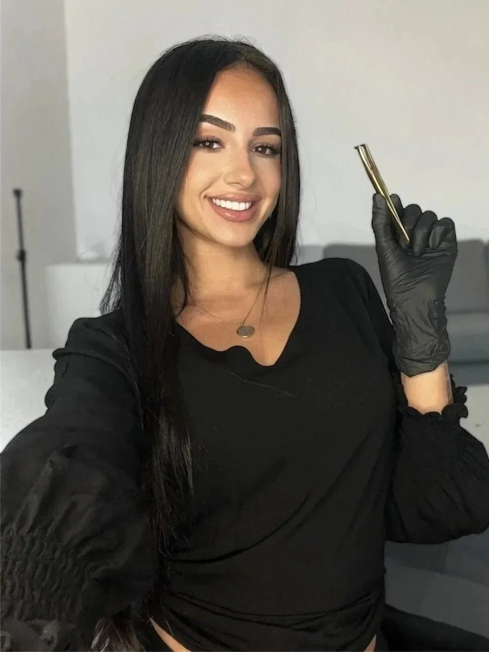 A woman with long dark hair smiling, holding a gold pen in black-gloved hand, sitting in a modern room with neutral decor.