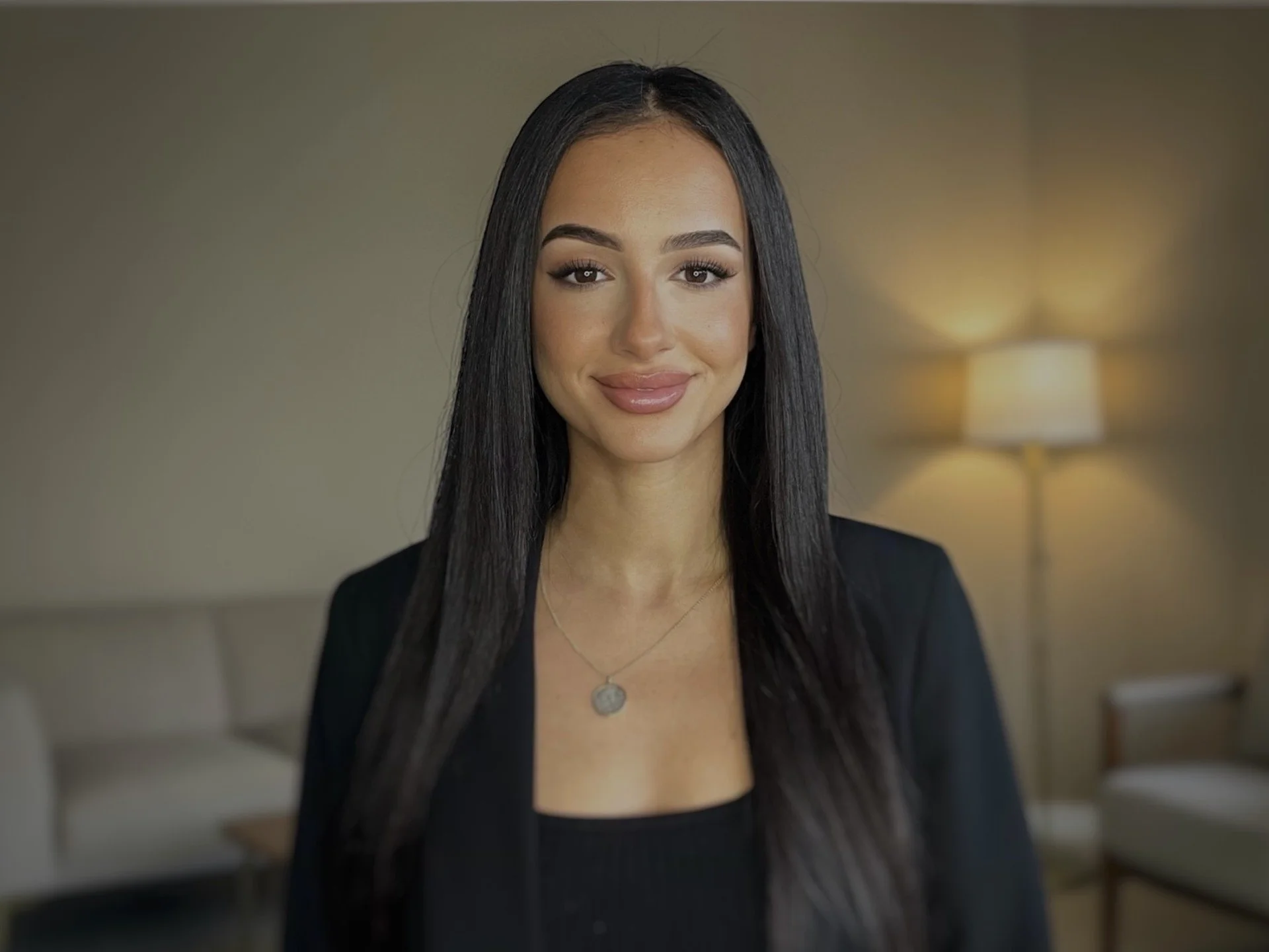 A woman with long black hair and light makeup smiling, wearing a black top and blazer, with a necklace, standing in a softly lit room with a beige wall, a sofa, and a lampshade in the background.