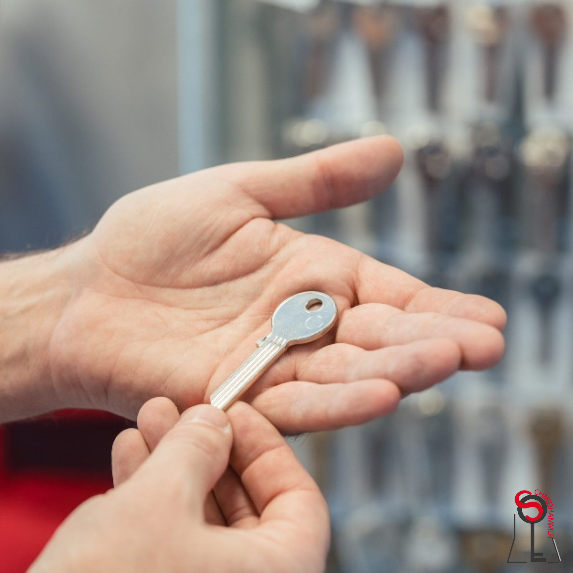 A person handing a silver key with a round head to another person's open palm. The background shows a blurred view of multiple similar keys hanging on a display.