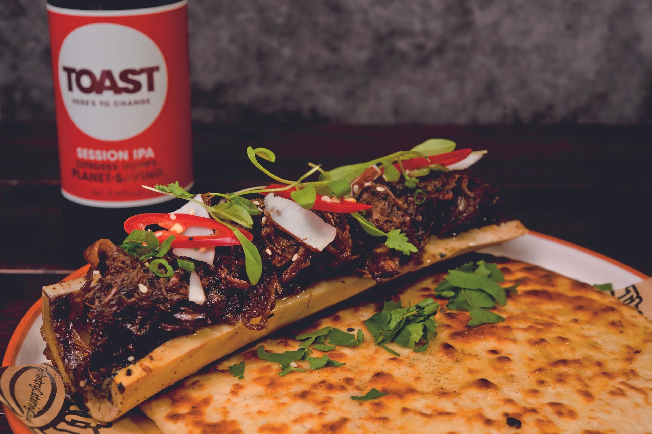 A plate with a toasted flatbread topped with braised beef, garnished with microgreens, sliced red chili, and coconut flakes. A piece of cheese or additional flatbread is on the side. In the background, there is a can of Toast Session IPA beer.