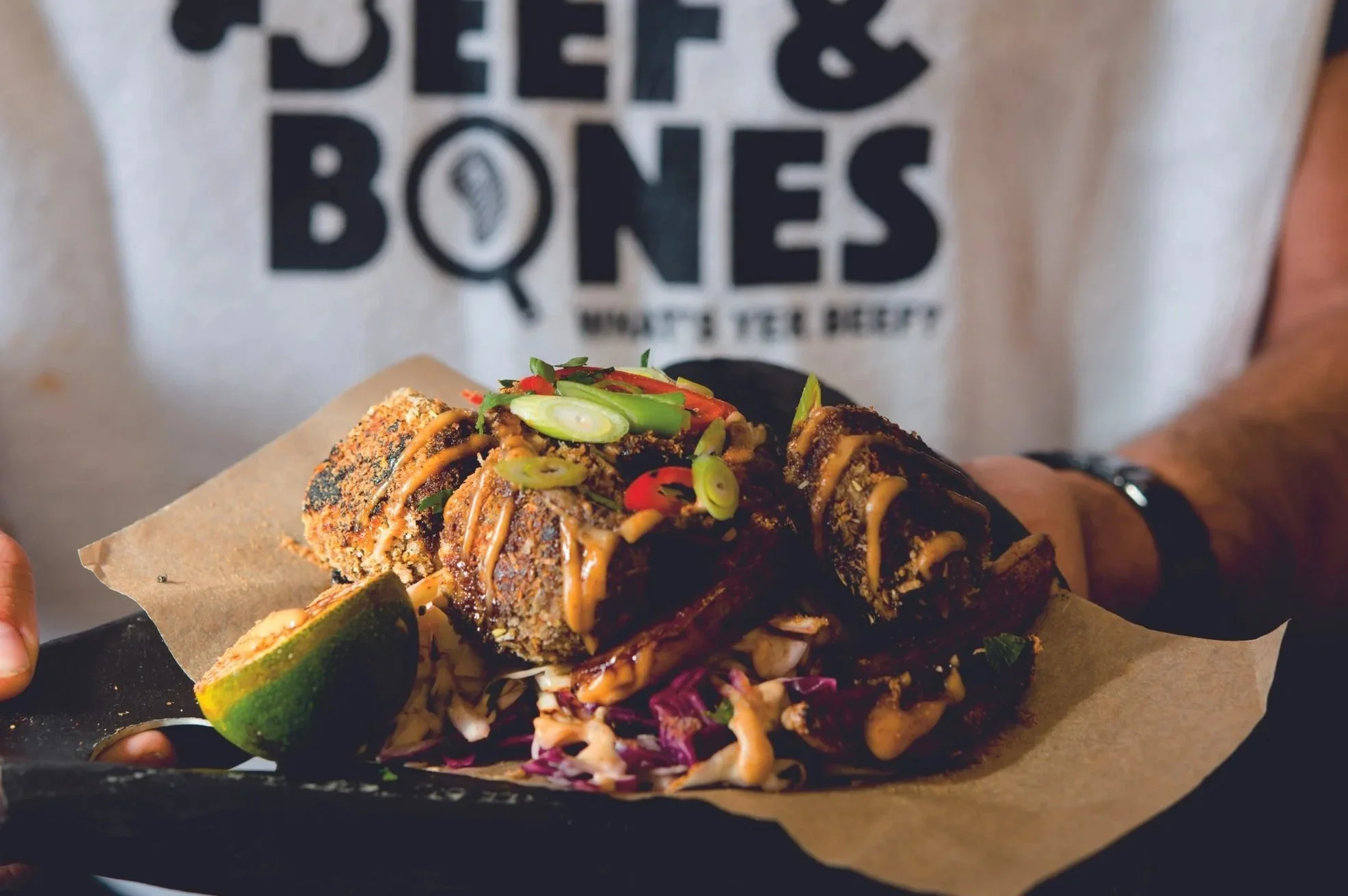 Fried food with sauce and green onion garnishes on a black tray with a lime wedge, held by a person wearing a white shirt with black text.