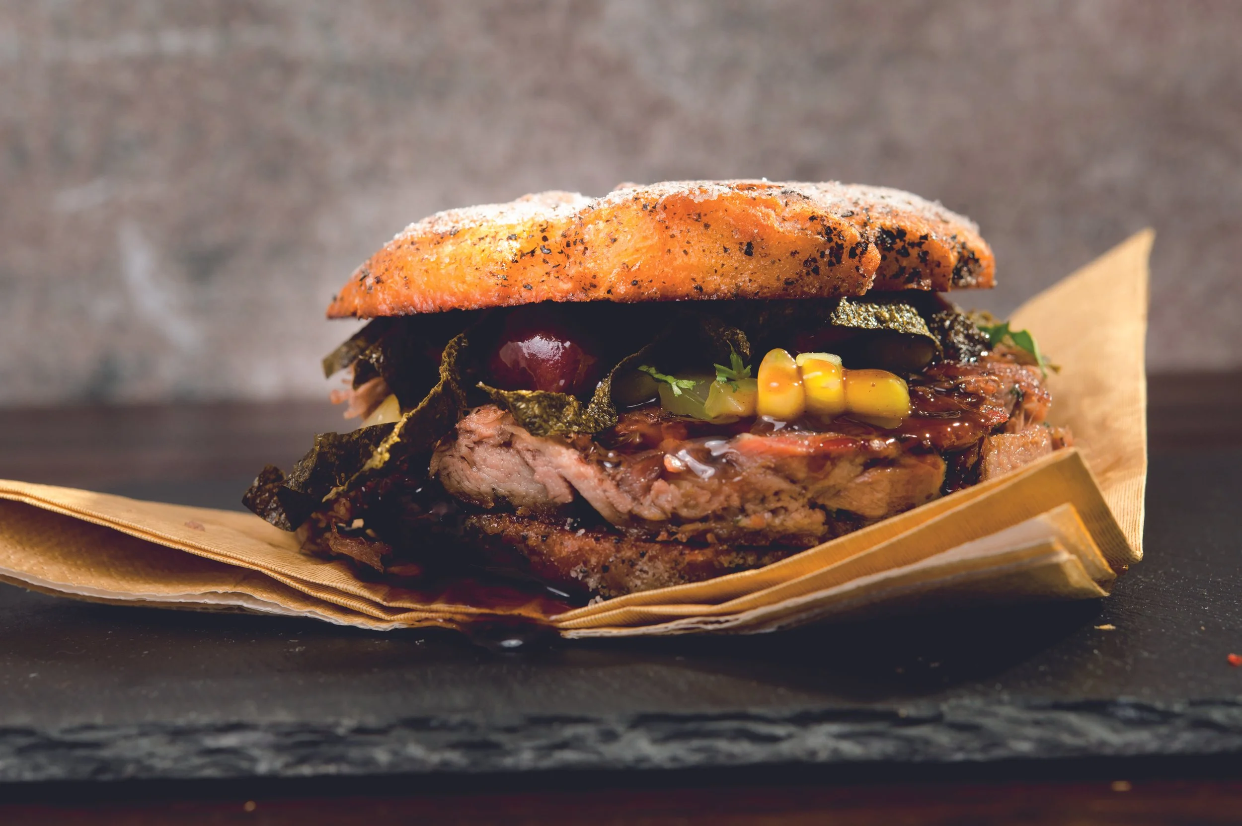 Close-up of a gourmet burger with grilled meat, corn, grapes, greens, and sauce, served on a paper-lined slate surface.