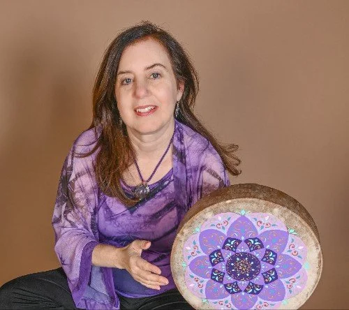 Woman with shoulder-length brown hair wearing a purple tie-dye shirt, sitting next to a large circular artwork with intricate purple, blue, and pink mandala design.