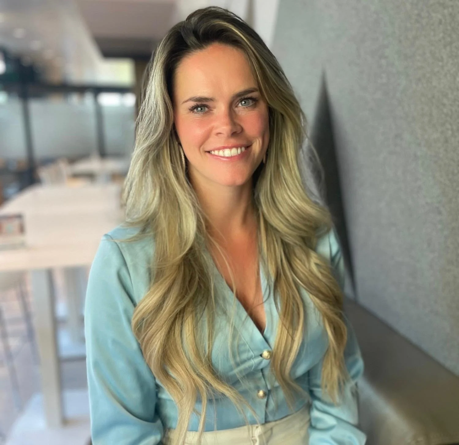 Portret van een lachende vrouw met lang, golvend blond haar, zittend in een kantoor met een grijze muur op de achtergrond.