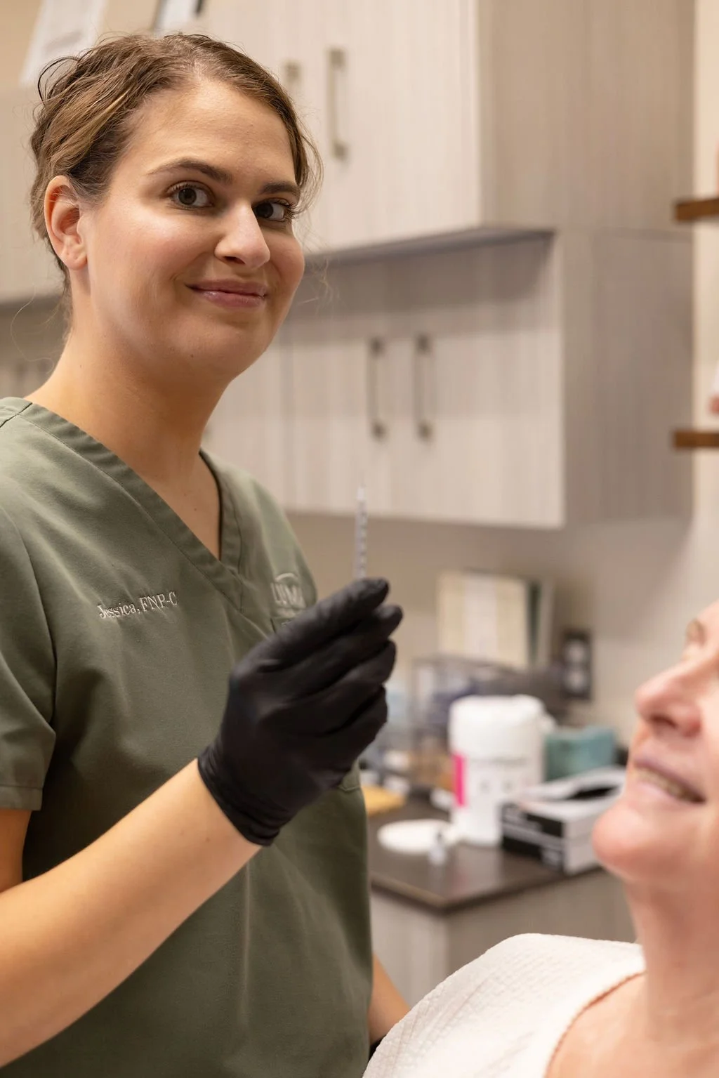 A smiling woman in medical scrubs holding a syringe in a clinical setting, with a patient's face partially visible.