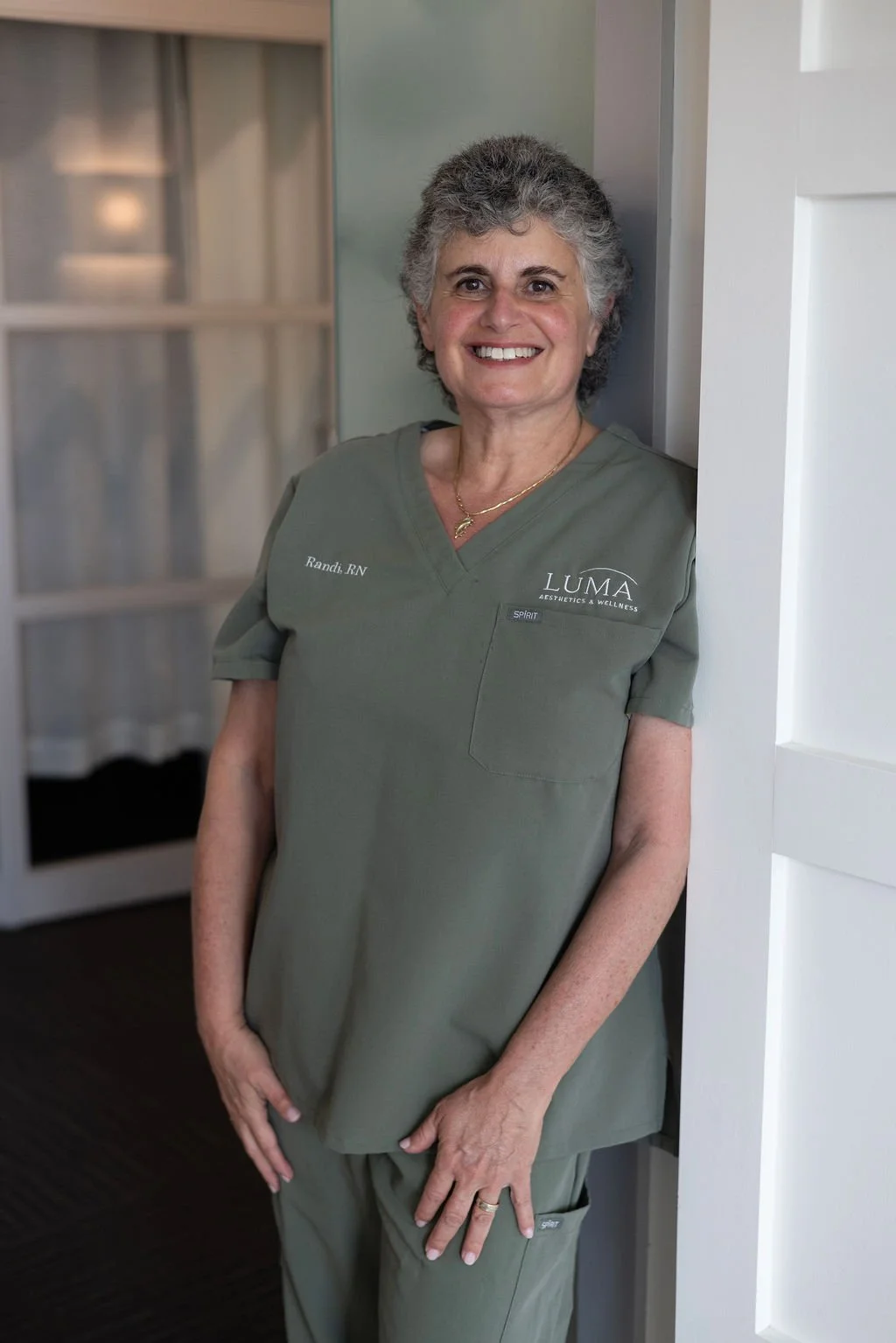 A smiling woman with gray hair wearing green medical scrubs standing and leaning against a wall in a room.