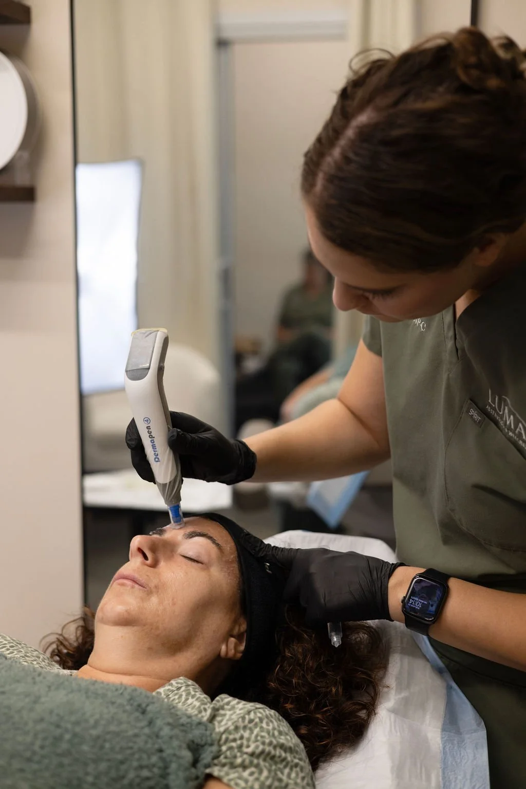 A woman lying on a medical bed receiving a cosmetic facial treatment with a handheld device, while a skincare professional in black gloves operates the device and wears a smartwatch.