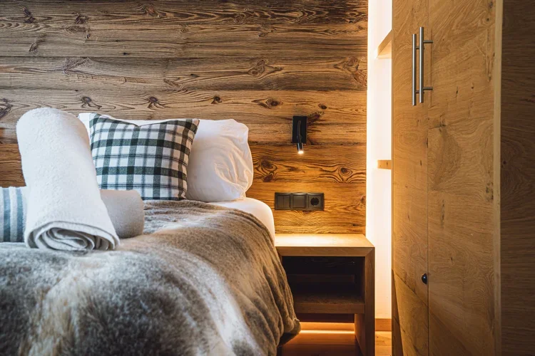 A cozy bedroom with a wooden wall, a bed with white and plaid pillows, a rolled towel, and a bedside table with a lamp.