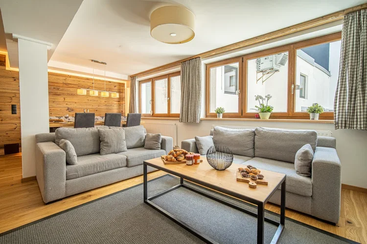 Living room with two gray sofas, a wooden coffee table with snacks and a vase, large windows with curtains, and a wooden floor.
