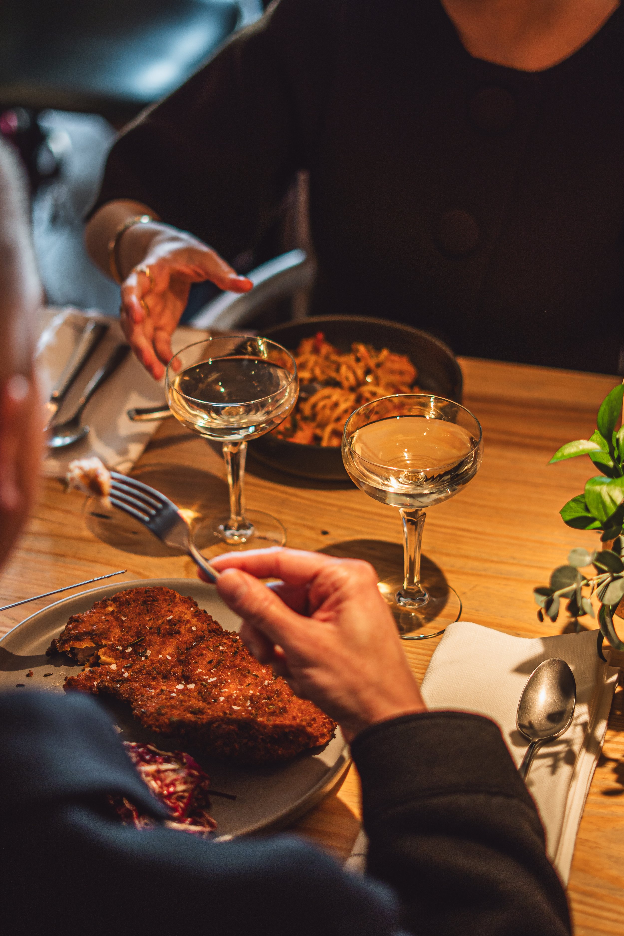 People dining at a restaurant with a plate of breaded fried fish, glasses of white wine, and a pasta dish on the table, with a small plant and cutlery.