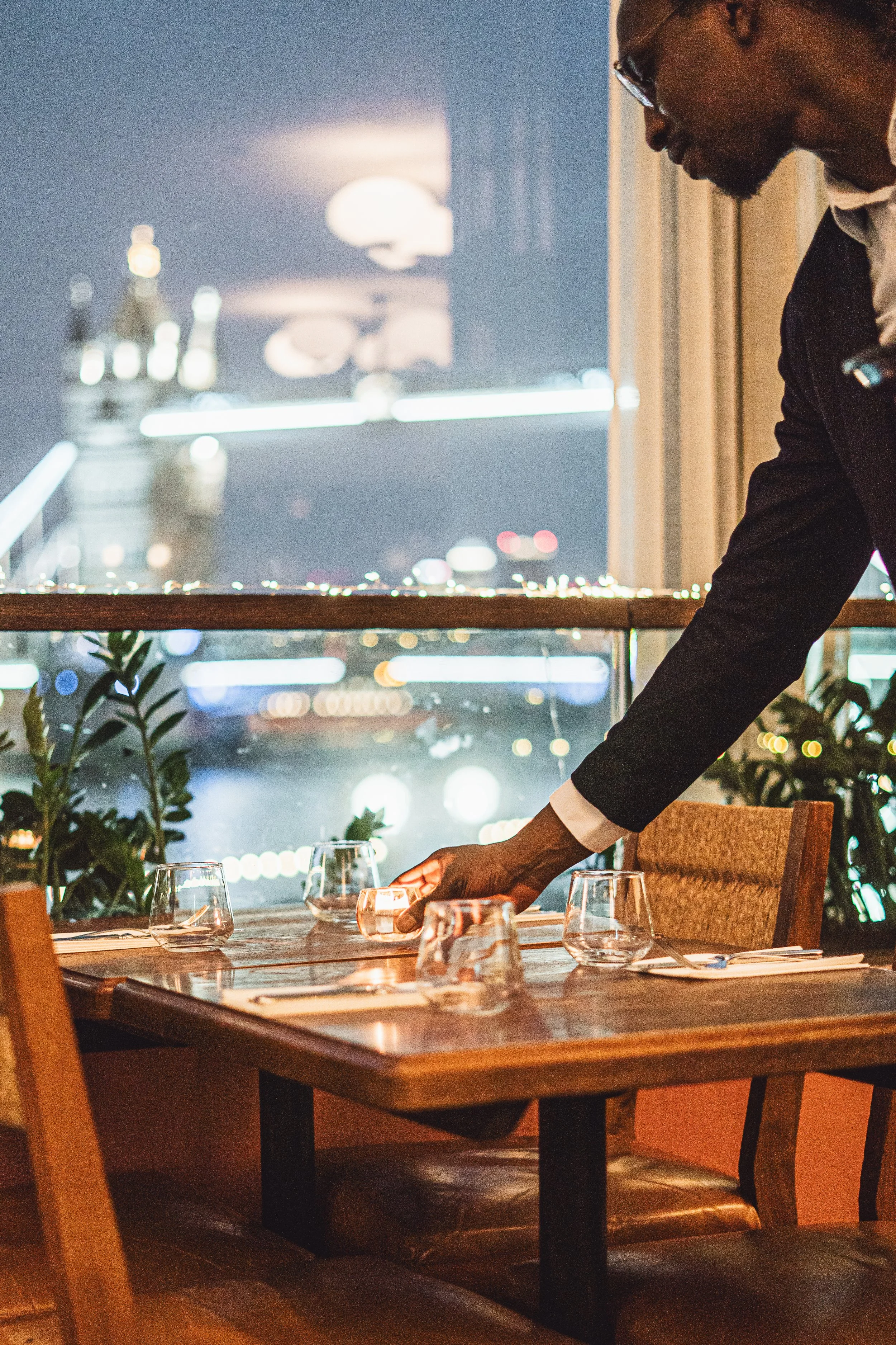 A server setting a table in a restaurant with a cityscape view at night in the background.