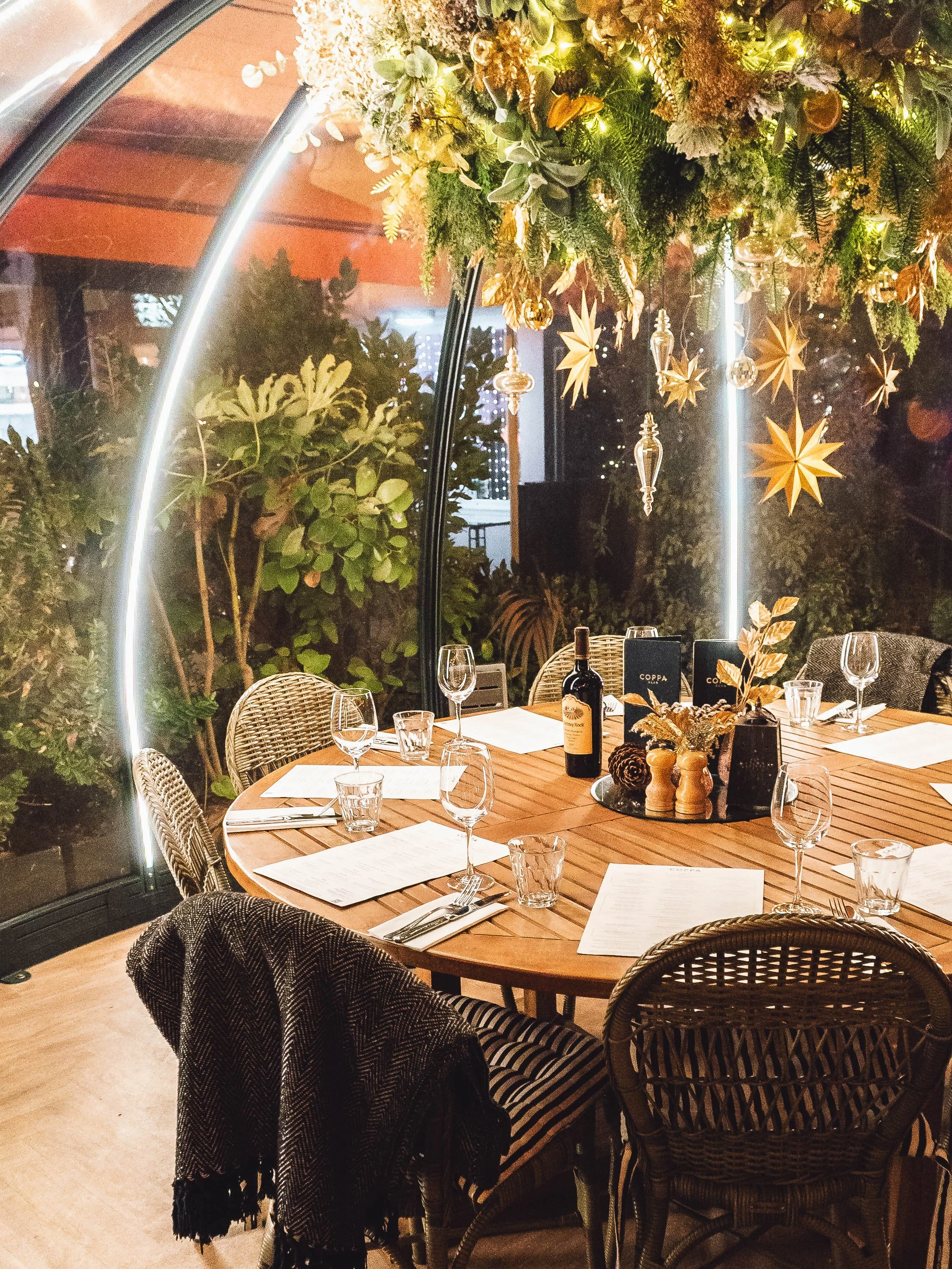 A decorated dining table inside a greenhouse-style space, with hanging gold star and ornament decorations, a floral centerpiece, wine glasses, menus, and a bottle of wine, illuminated by warm lighting.